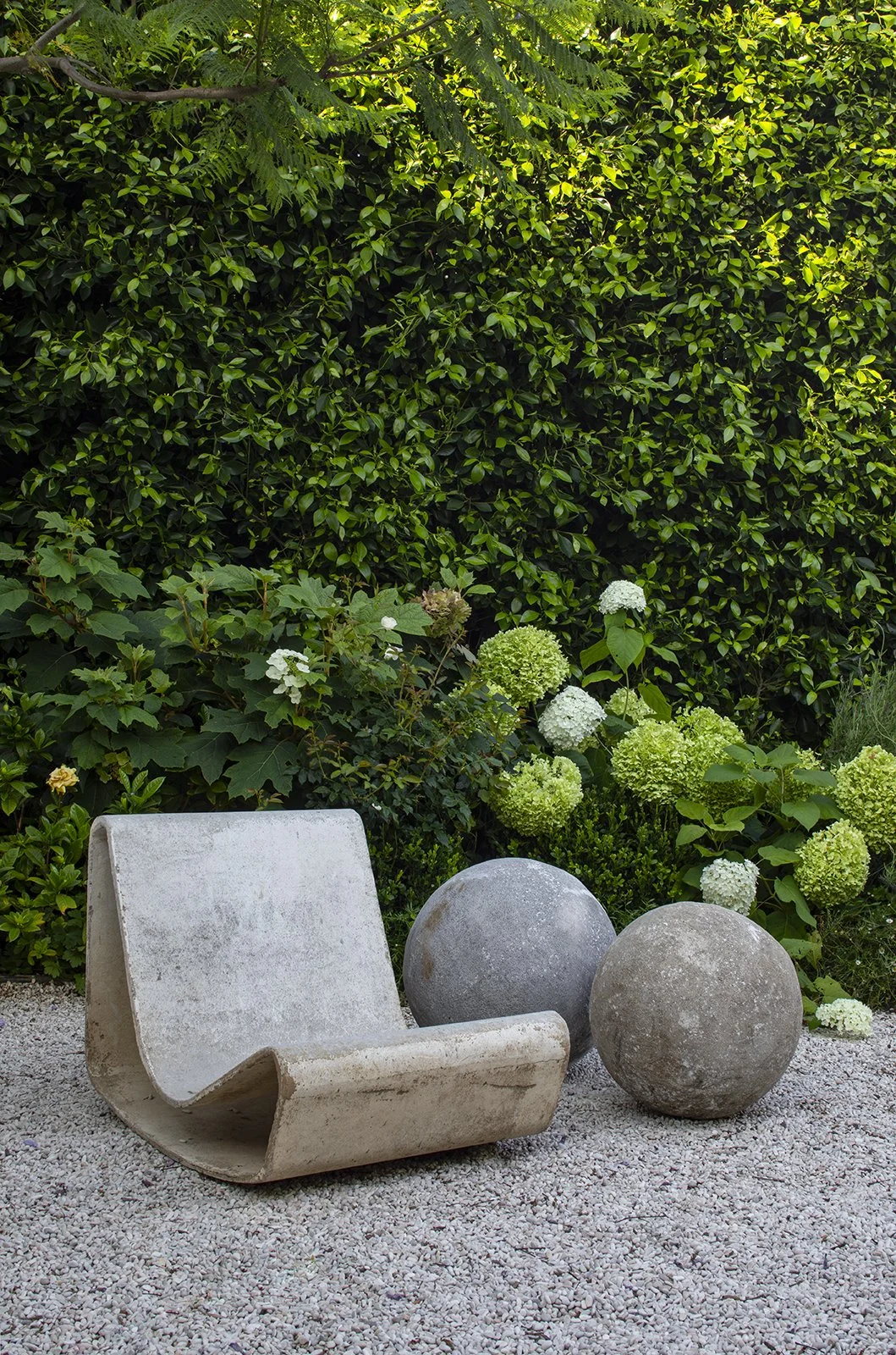 Modern outdoor garden scene with a concrete chair and two large spherical stone sculptures, set against lush green shrubbery and blooming hydrangeas.