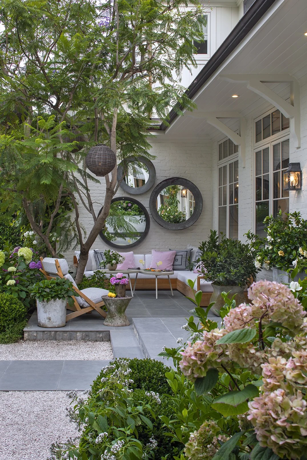 A cozy outdoor patio space featuring a corner seating area with cushions, surrounded by lush green plants and flowers, with decorative round mirrors on the white brick wall.