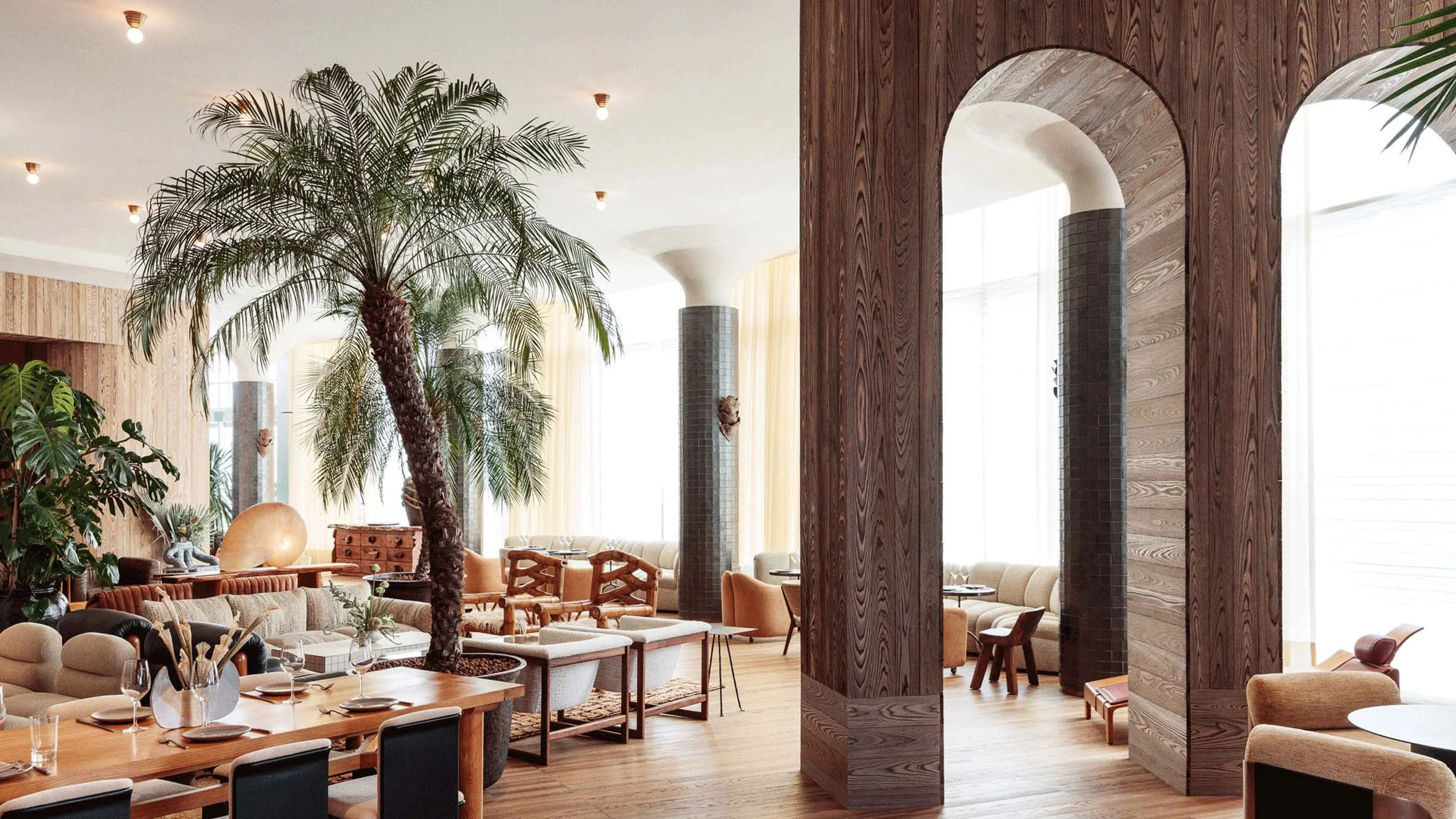 Interior of a modern restaurant with wooden accents, large windows, a tall palm tree, and various seating areas including tables and chairs.