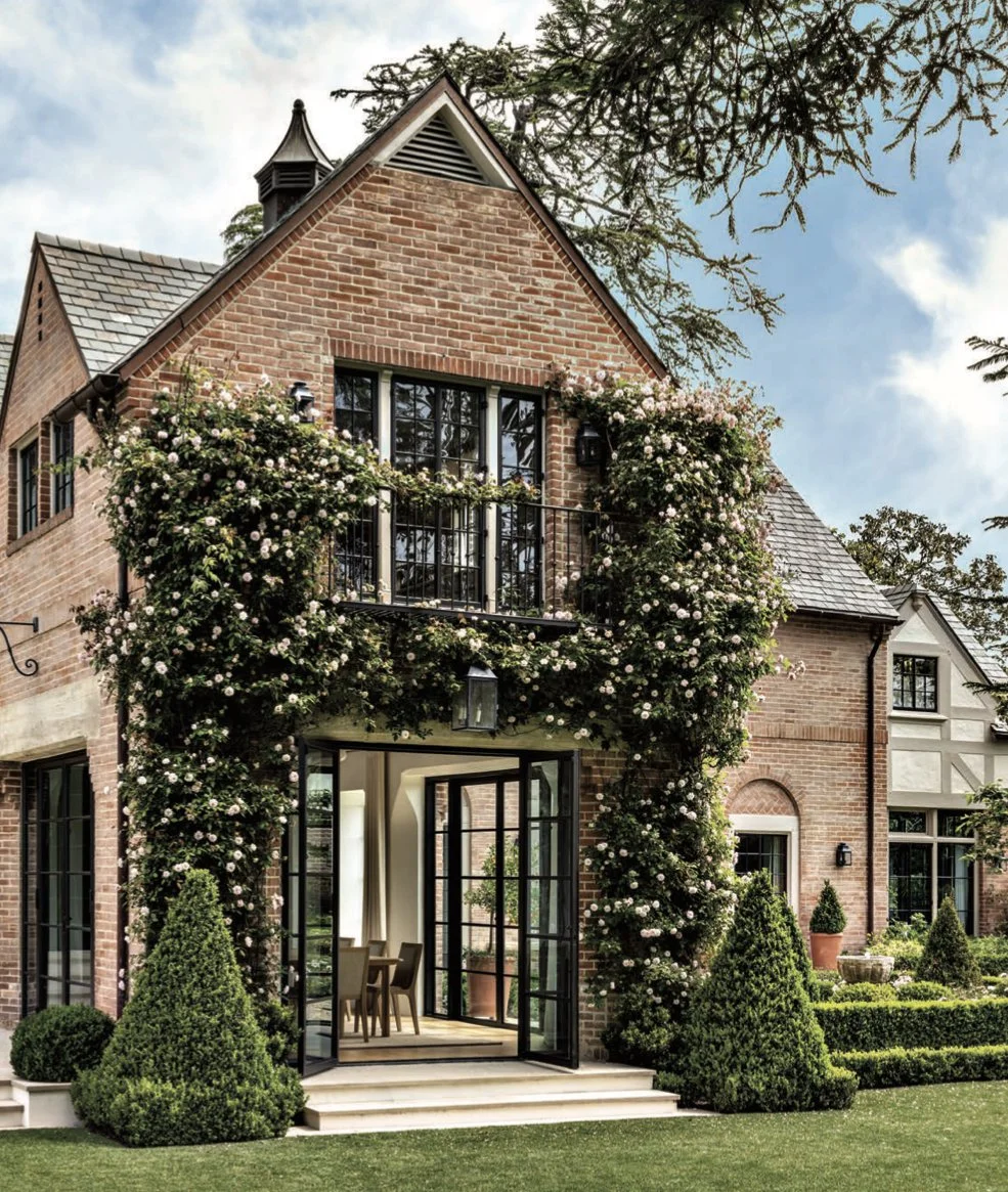 A beautiful brick house with a lush garden and climbing roses around the entrance, large glass doors leading into the interior, and a balcony on the upper floor.