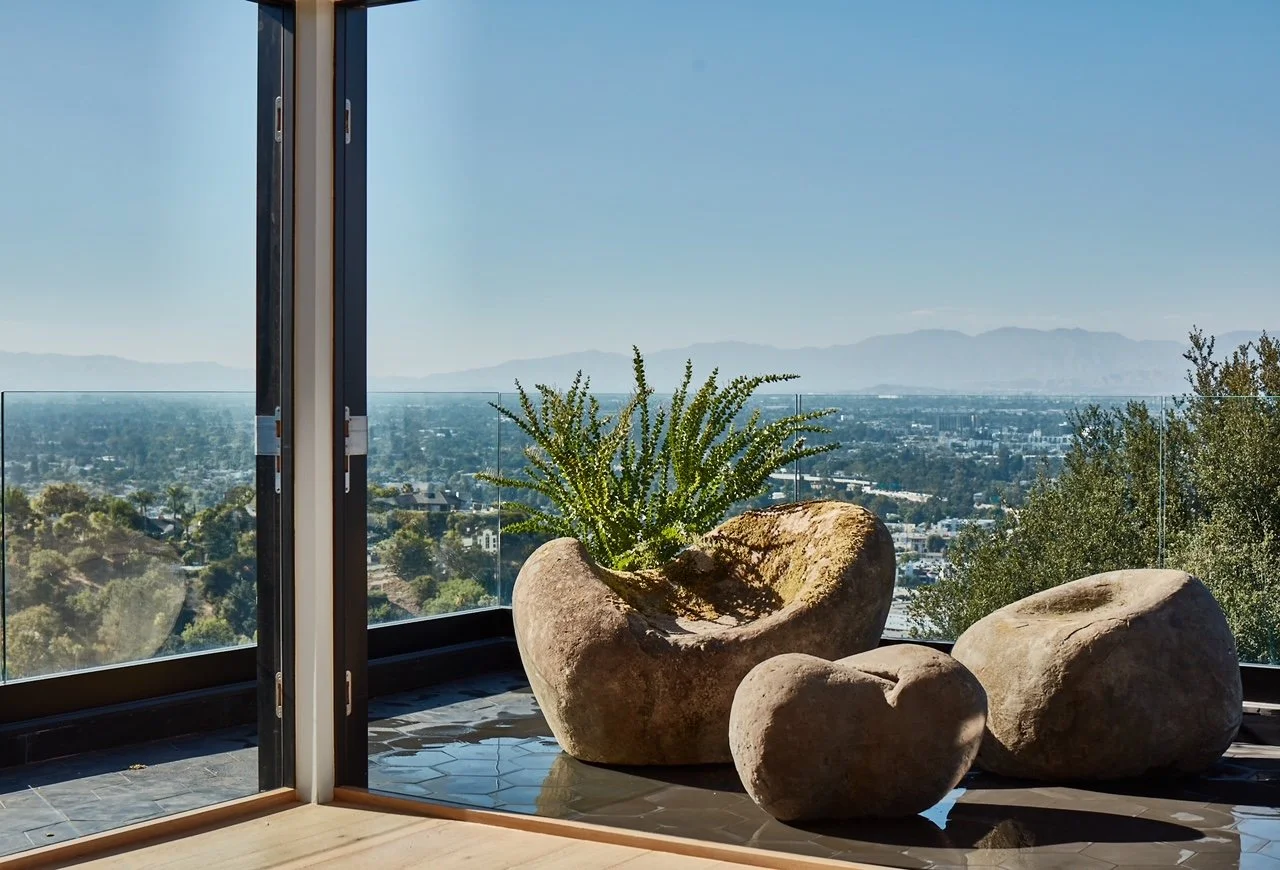Interior with three large rocks and a potted cactus near a glass railing, overlooking a cityscape and mountains under a blue sky.