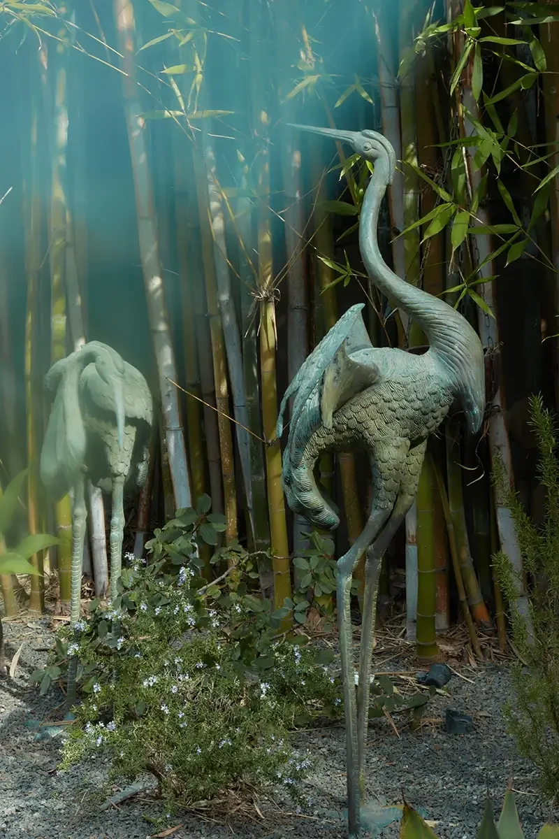 Two metal sculptures of herons or cranes are placed among bamboo plants and other greenery in a garden setting.