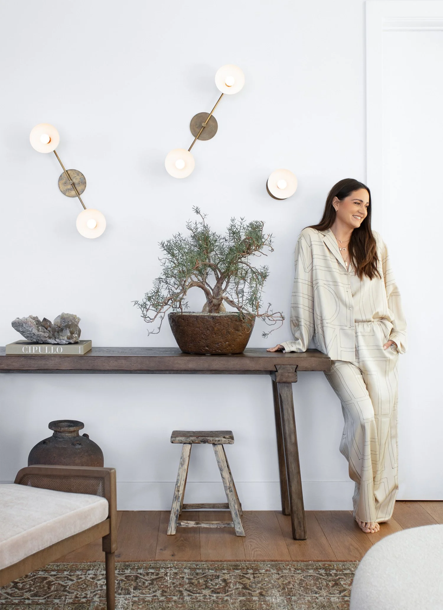 A woman in cream-colored pajamas standing and leaning against a wooden table with potted plant, decorative rock, and book in a bright, modern living room with artistic lighting on the wall.