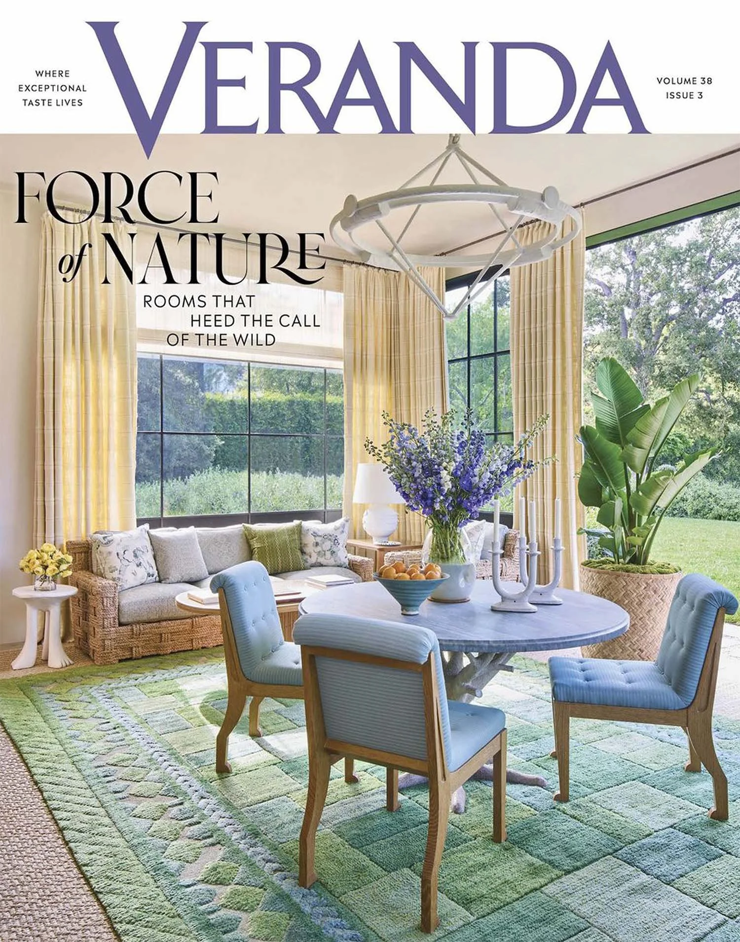 Cover of Veranda magazine featuring a bright, sunlit living room with large windows, a wicker sofa with cushions, a round marble dining table with four upholstered chairs, and green plants. A floral centerpiece and decorative candle holders are on the table, and the room has yellow curtains and a green patterned rug.