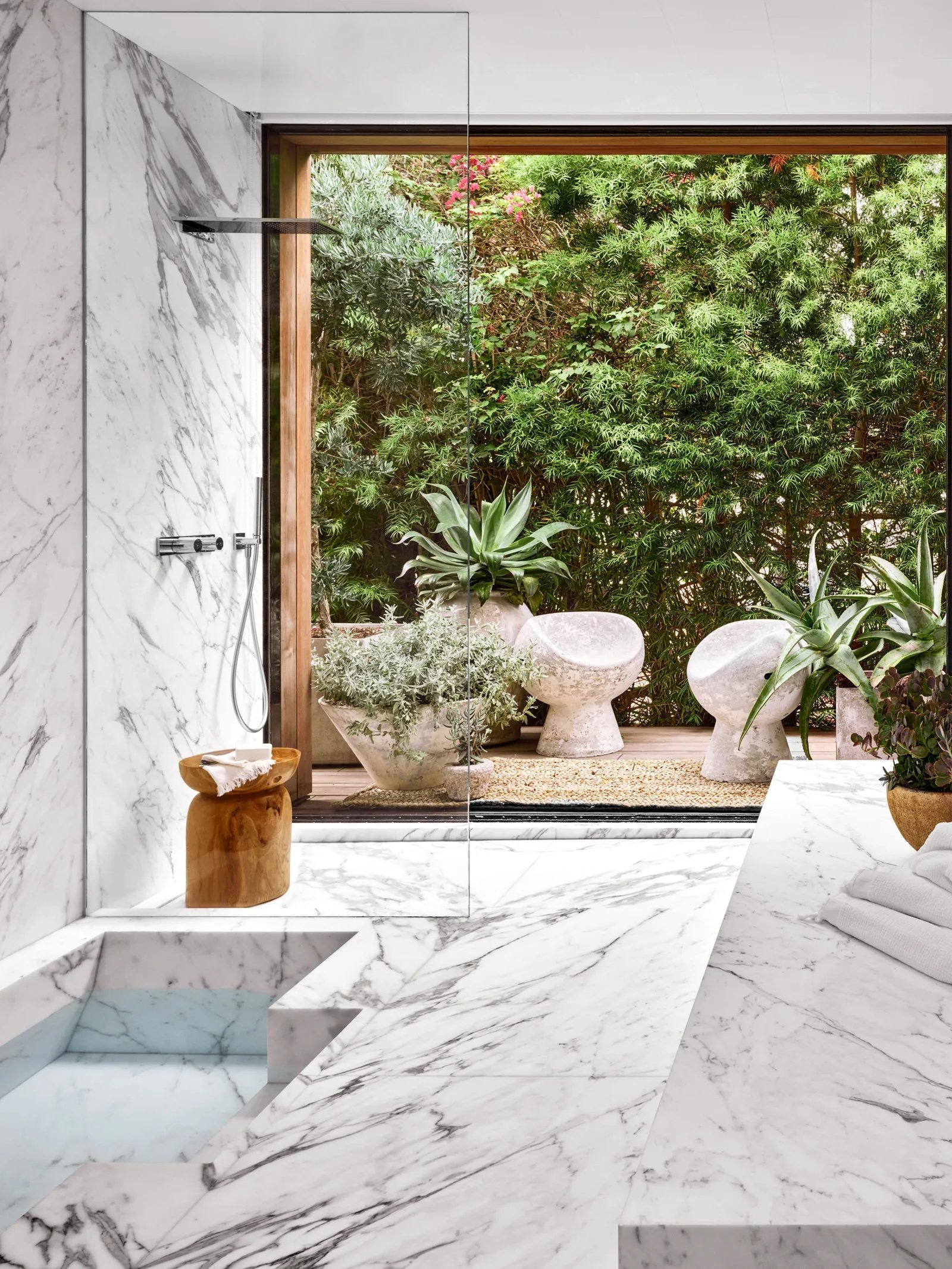 Luxurious bathroom with white marble surfaces and a large glass door opening to a lush garden with potted plants and stone benches.