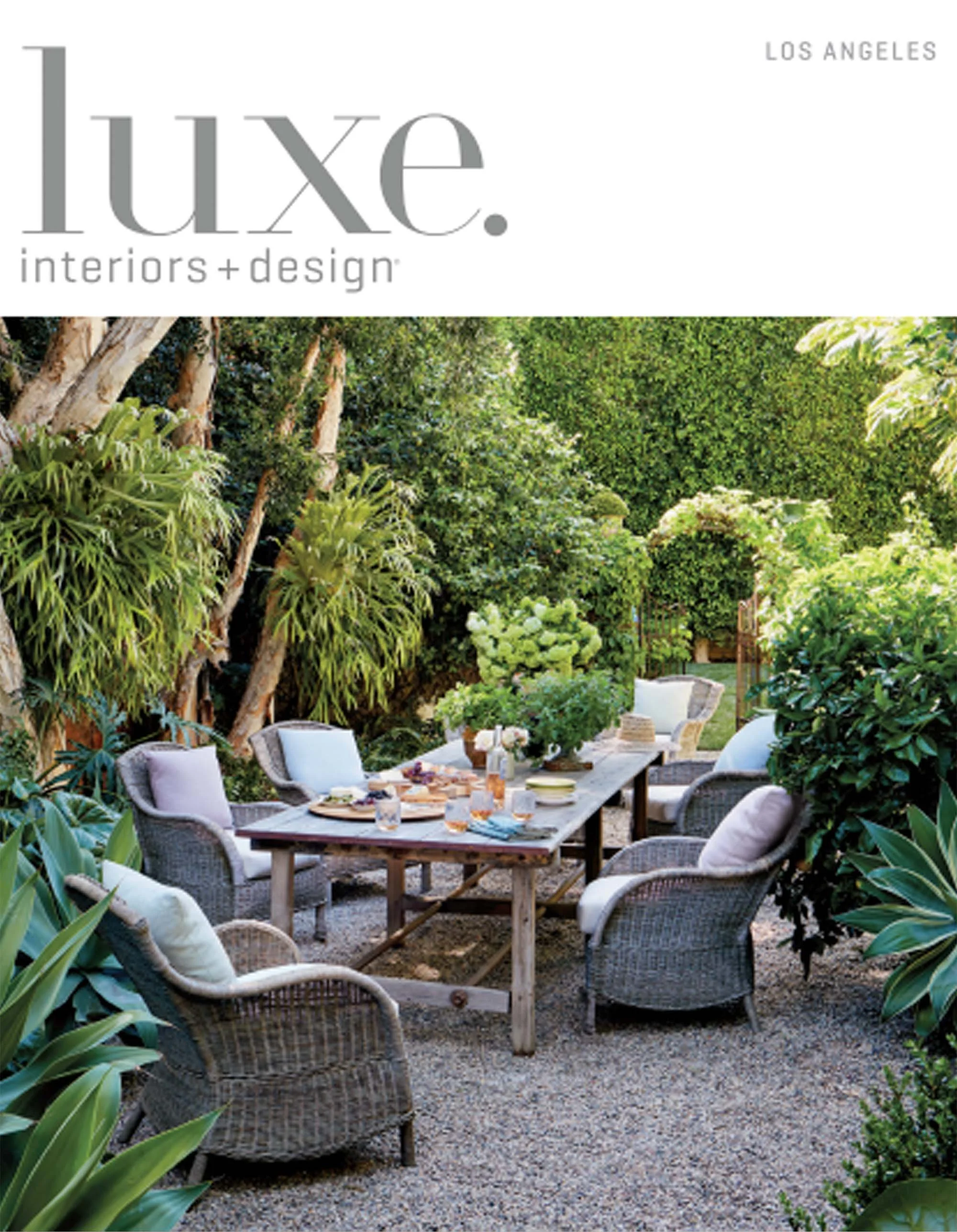 An outdoor garden dining area with six wicker chairs with cushions around a rustic wooden table set for a meal, surrounded by lush green trees and bushes.