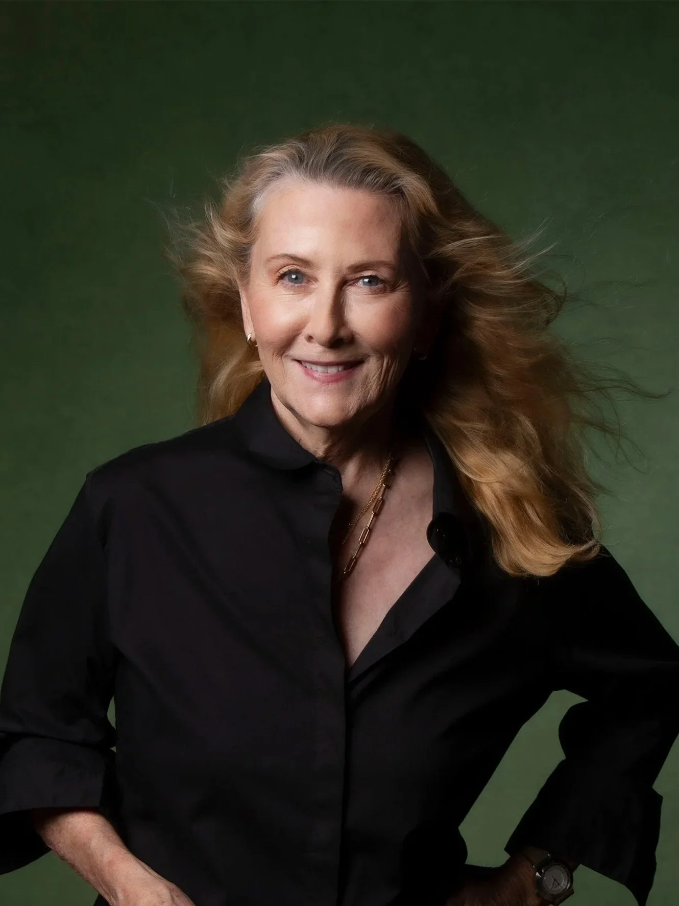 A middle-aged woman with long, wavy blonde hair smiling and dressed in a black button-down shirt, standing against a green background.