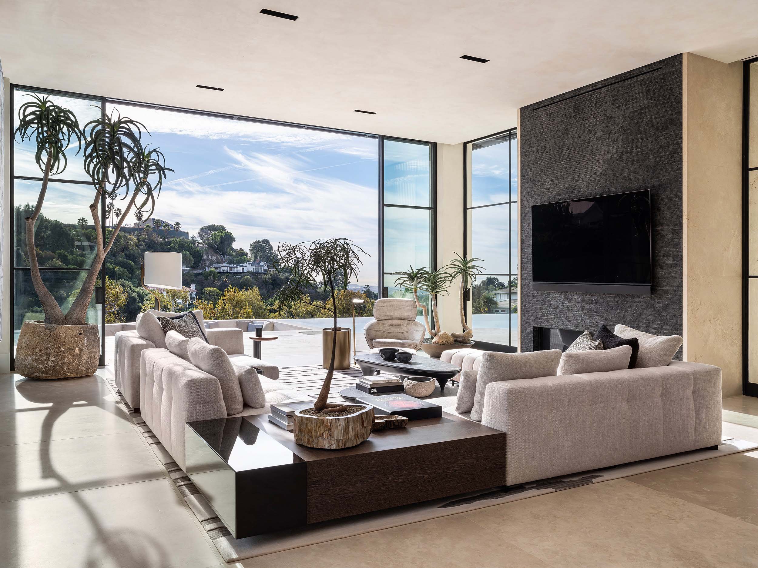 Modern living room with large floor-to-ceiling windows, white sofas, potted plants, and a black fireplace with a mounted television.