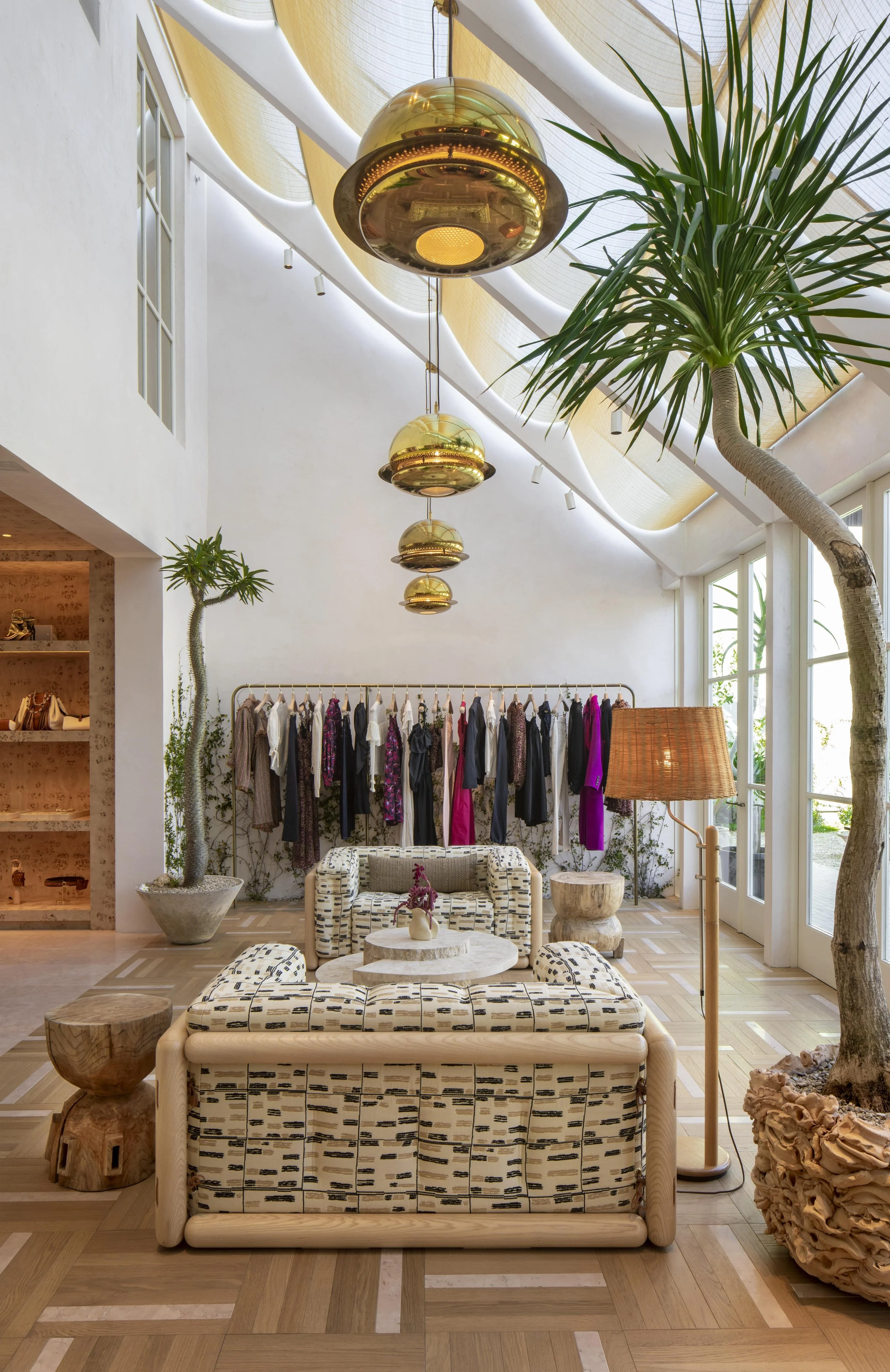 Interior of a boutique or showroom with hanging clothes, indoor plants, wooden furniture, and gold pendant lights