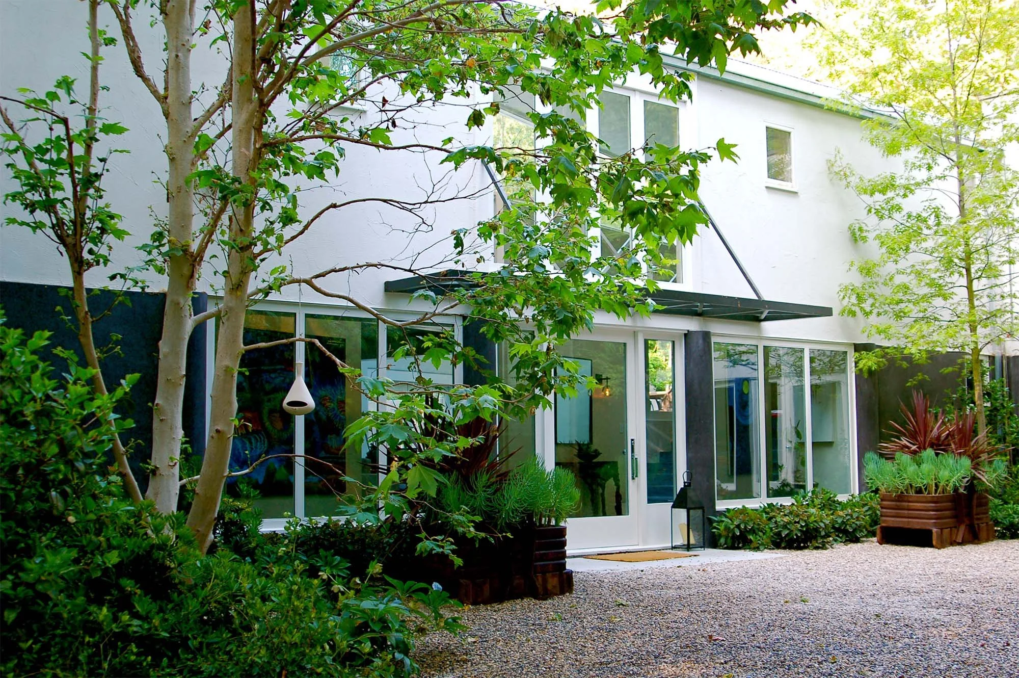 Home with a garden featuring trees, potted plants, and a gravel pathway, with large glass doors and windows.