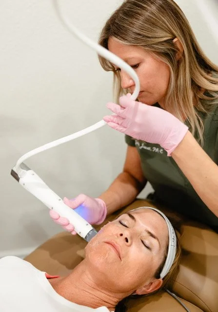 A woman with blonde hair in pink gloves is performing a skin treatment on a woman lying down with her eyes closed in a spa or clinic setting.