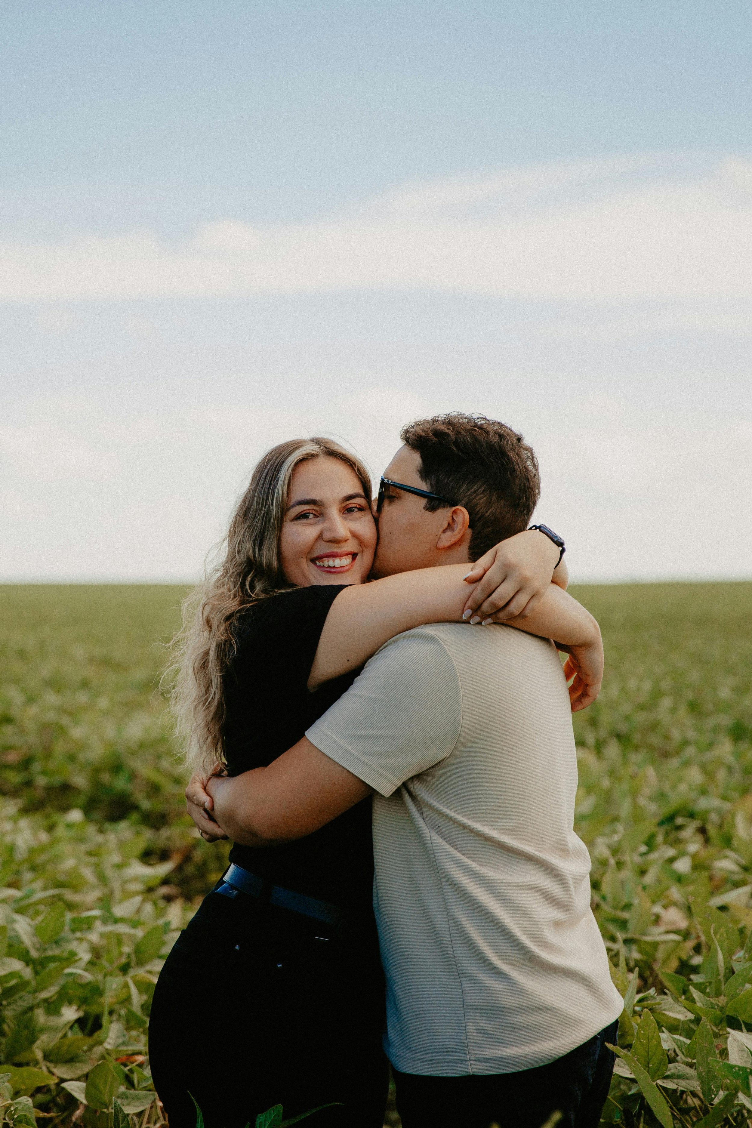 A couple hugging outdoors in a field, with the woman smiling at the camera and the man kissing her cheek.
