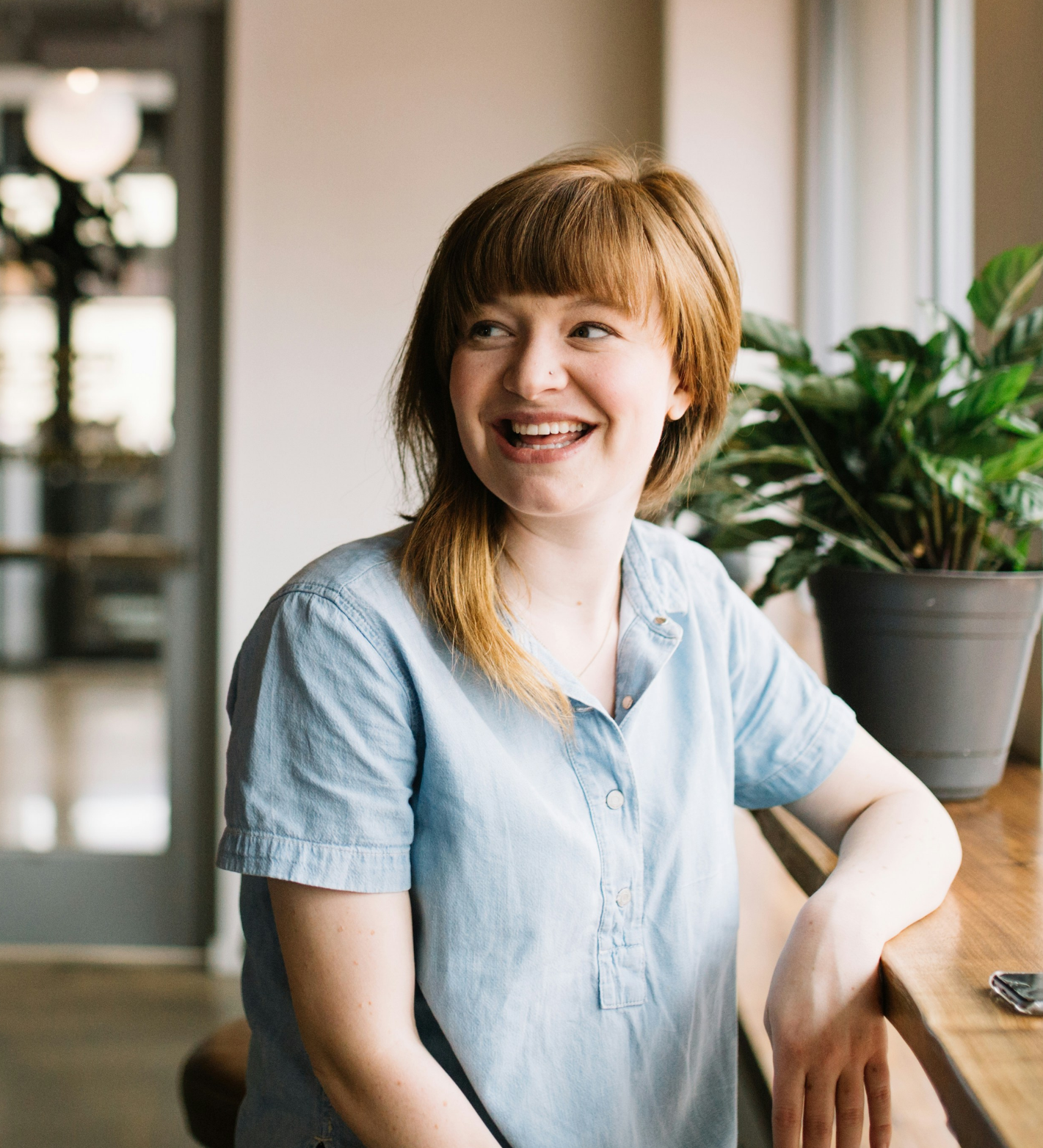 A young woman with red hair and a light blue shirt smiling while sitting at a wooden counter in a cozy cafe with a potted plant nearby.