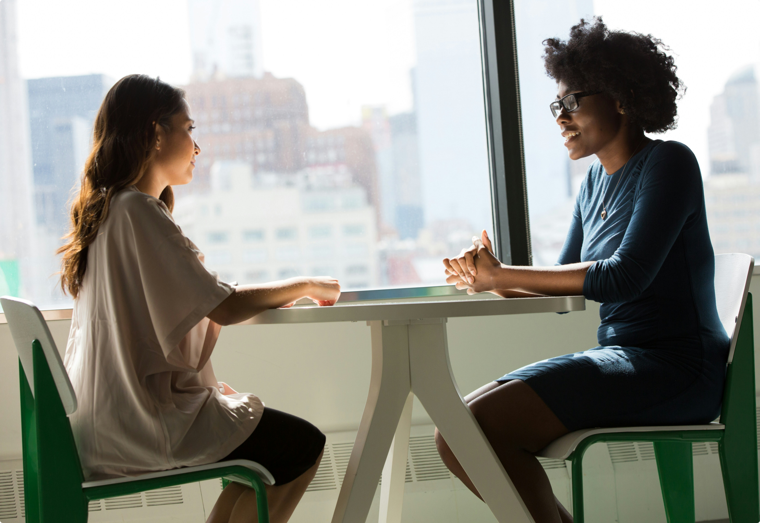 Two women sit at a table by a large window, having a conversation. The city skyline is visible outside.