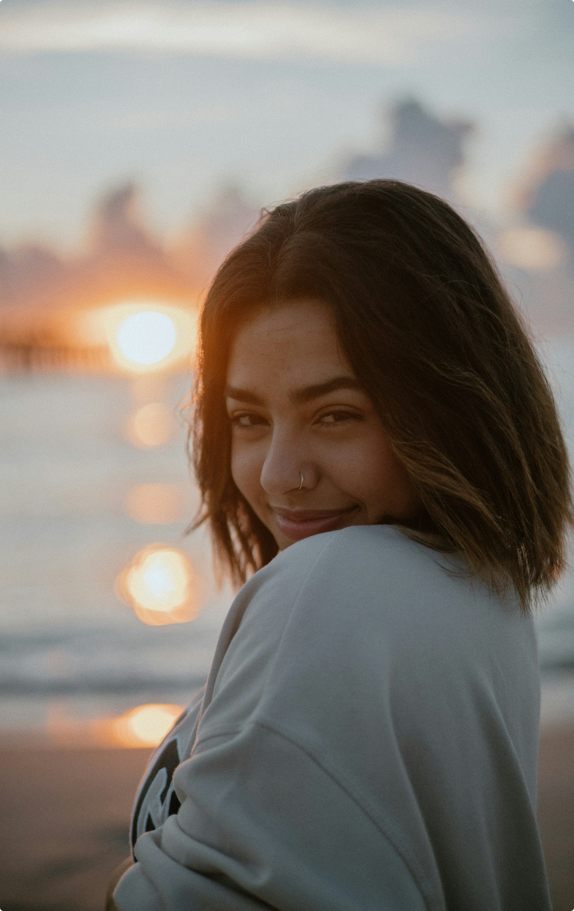 Young woman with shoulder-length brown hair, smiling and winking, at the beach during sunset with blurred clouds and water in the background.