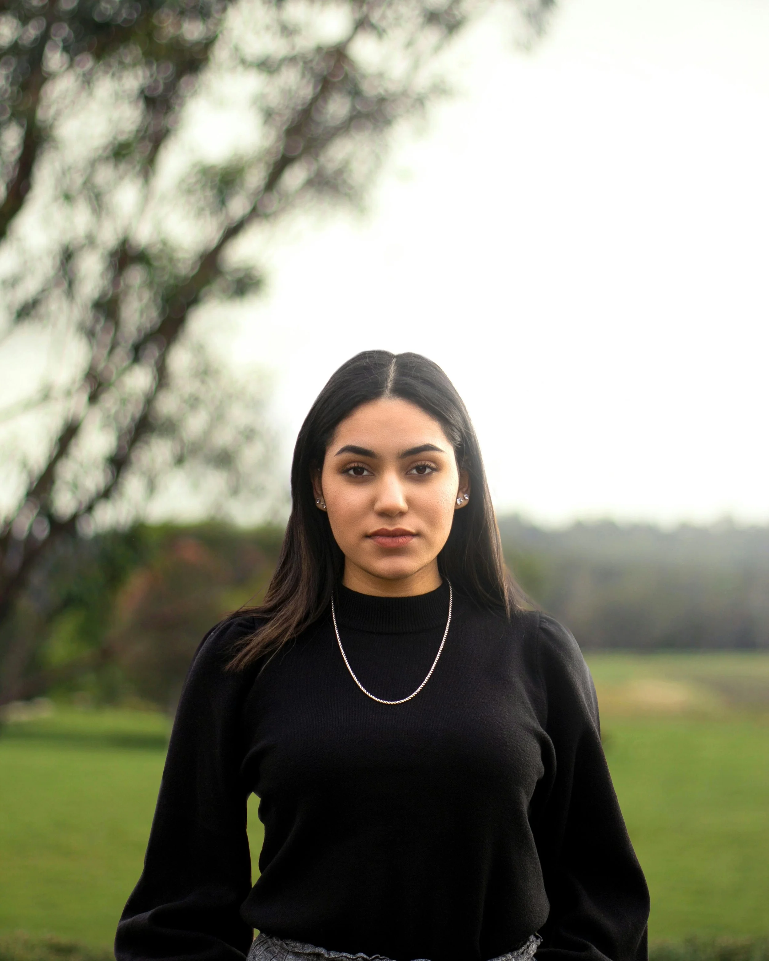 A young woman with dark hair and pierced ears stands outdoors in front of a tree and a grassy field on a cloudy day, wearing a black sweater and a silver necklace.