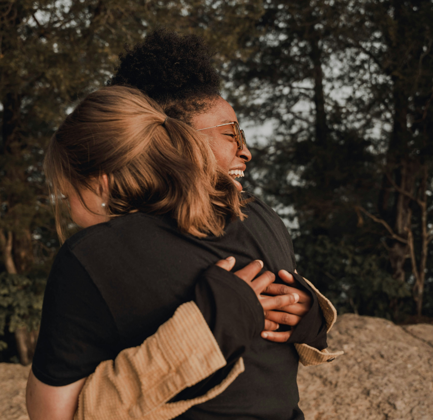 Two women embracing outdoors, smiling and joyful, with trees in the background during sunset.