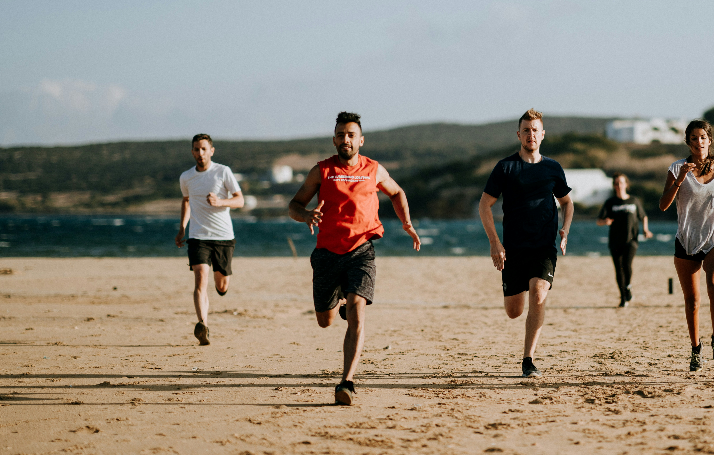 Group of five people running on a sandy beach near the ocean, with hills and some buildings in the background.