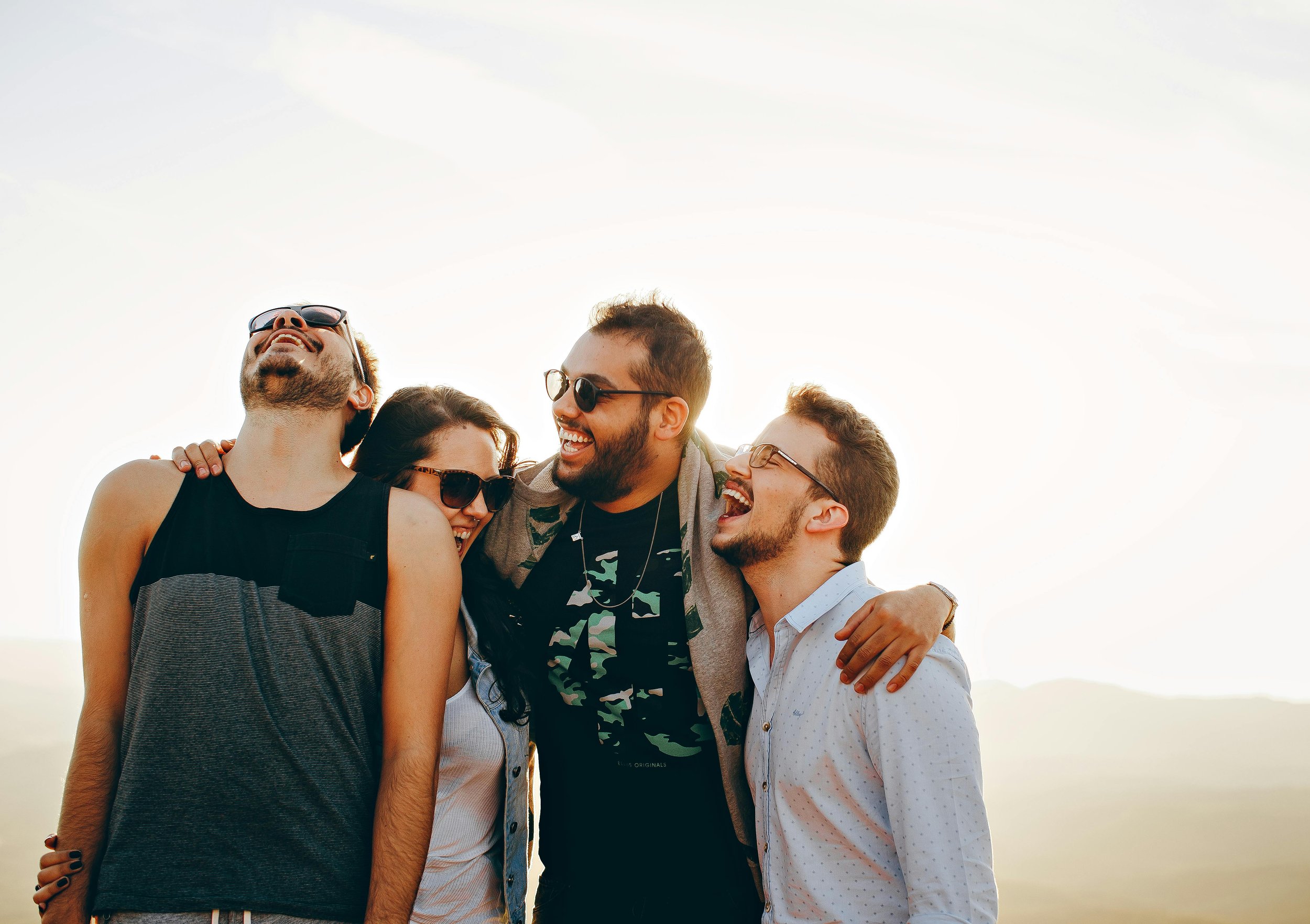Group of five friends smiling and laughing together outdoors during daytime, with arms around each other, wearing casual summer clothing and sunglasses, with a bright sky in the background.
