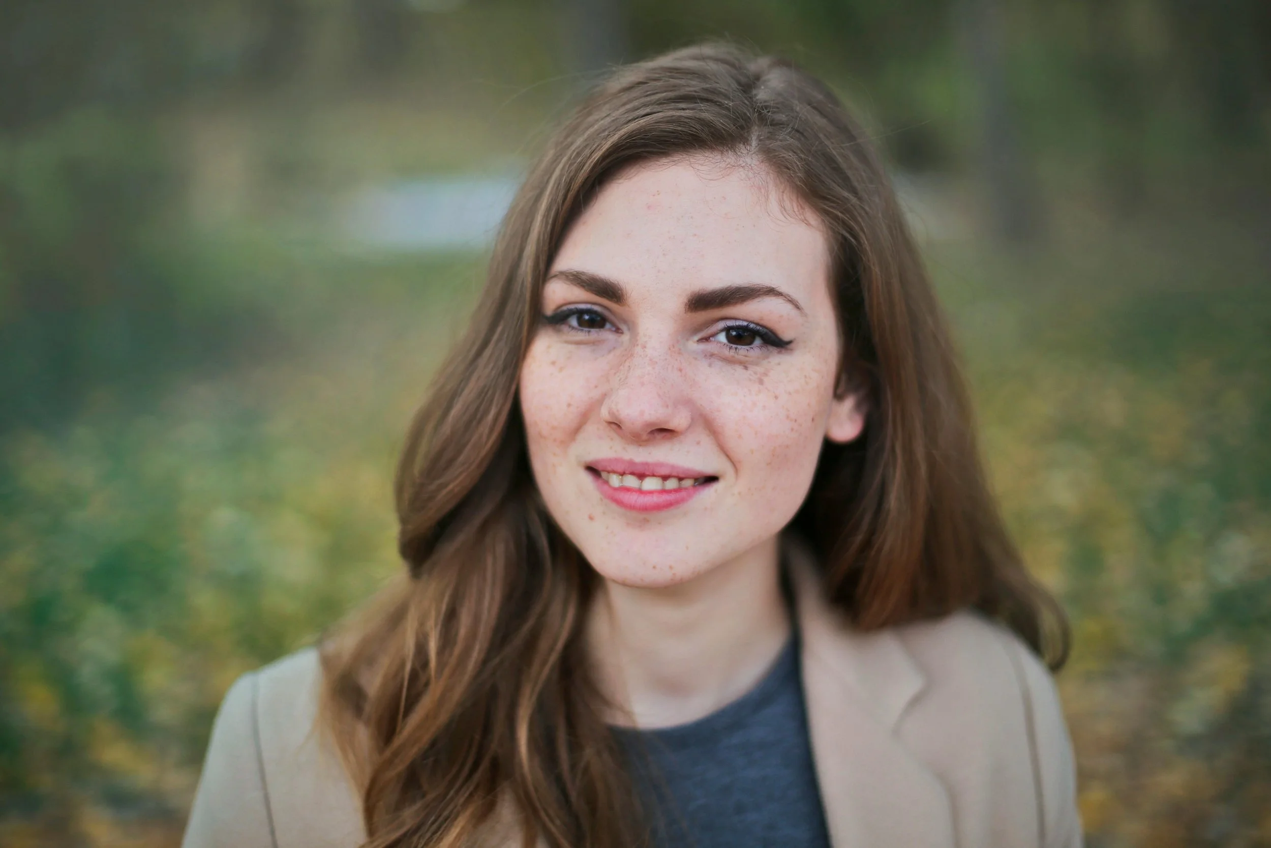 A young woman with long brown hair, freckles, and blue eyes smiling outdoors with blurred green and yellow foliage in the background.