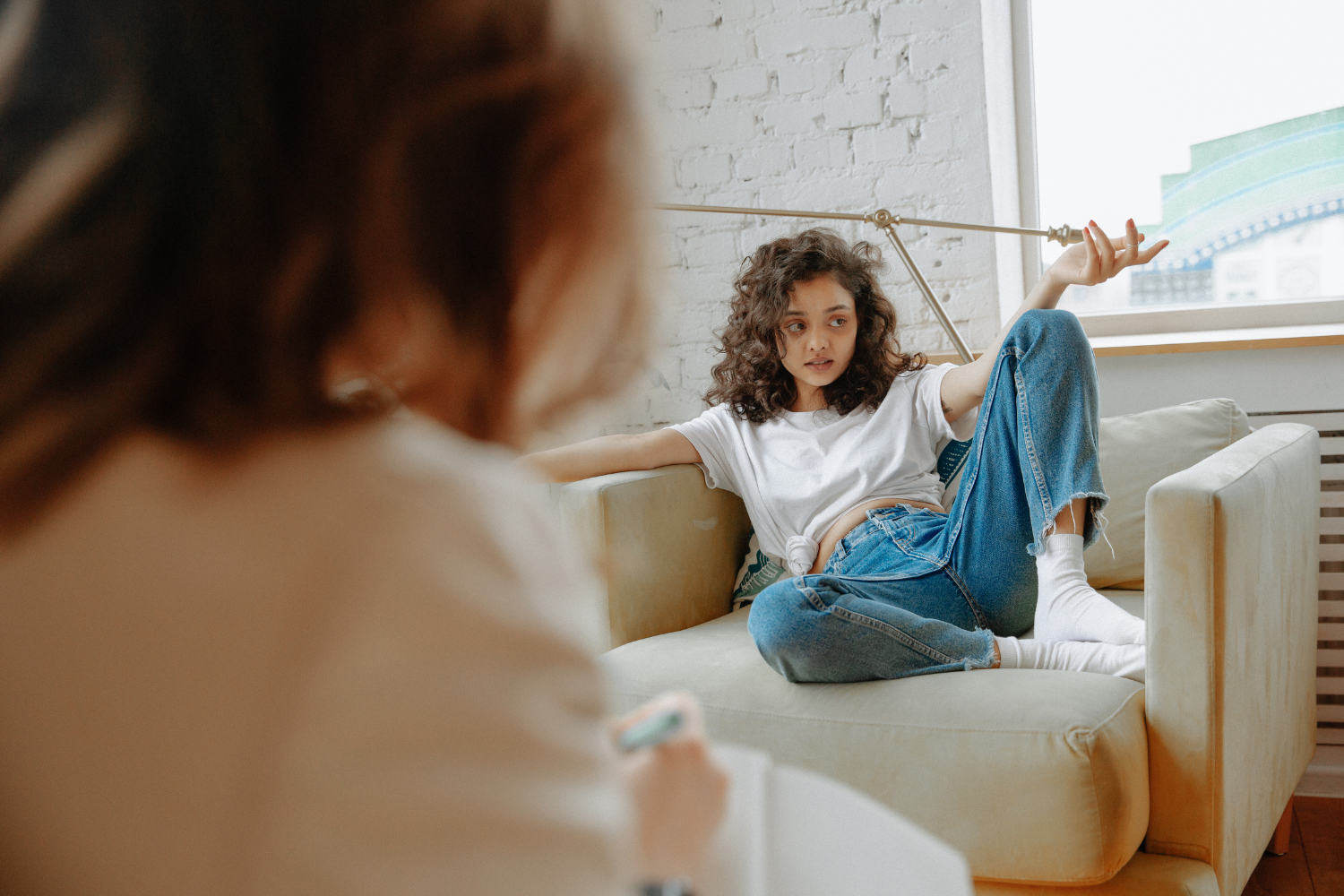A woman with red hair sitting on a couch, taking notes. A girl with curly hair in a white t-shirt and jeans is sitting on the couch, relaxed with one leg crossed over the other, gesturing with her hand while speaking.