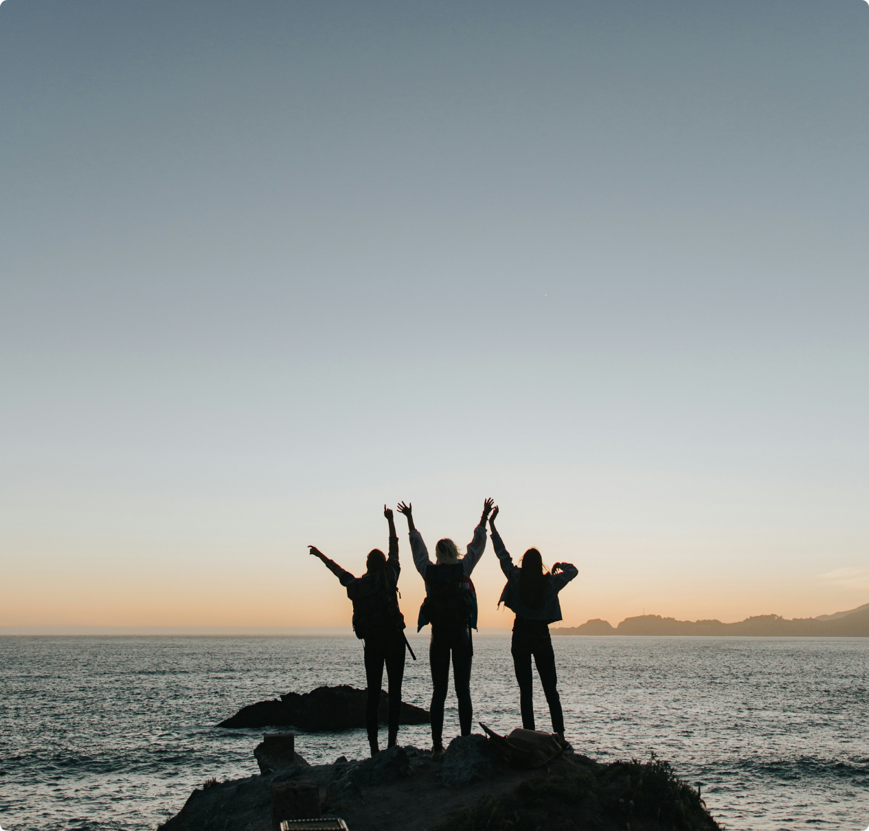 Three people standing on rocks by the ocean at sunset with arms raised in celebration.