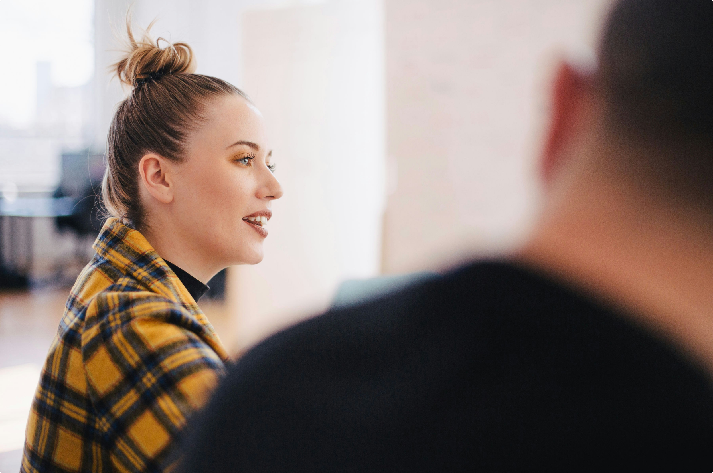 A woman with a high bun hairstyle is talking to someone in an office setting, wearing a yellow plaid blazer.