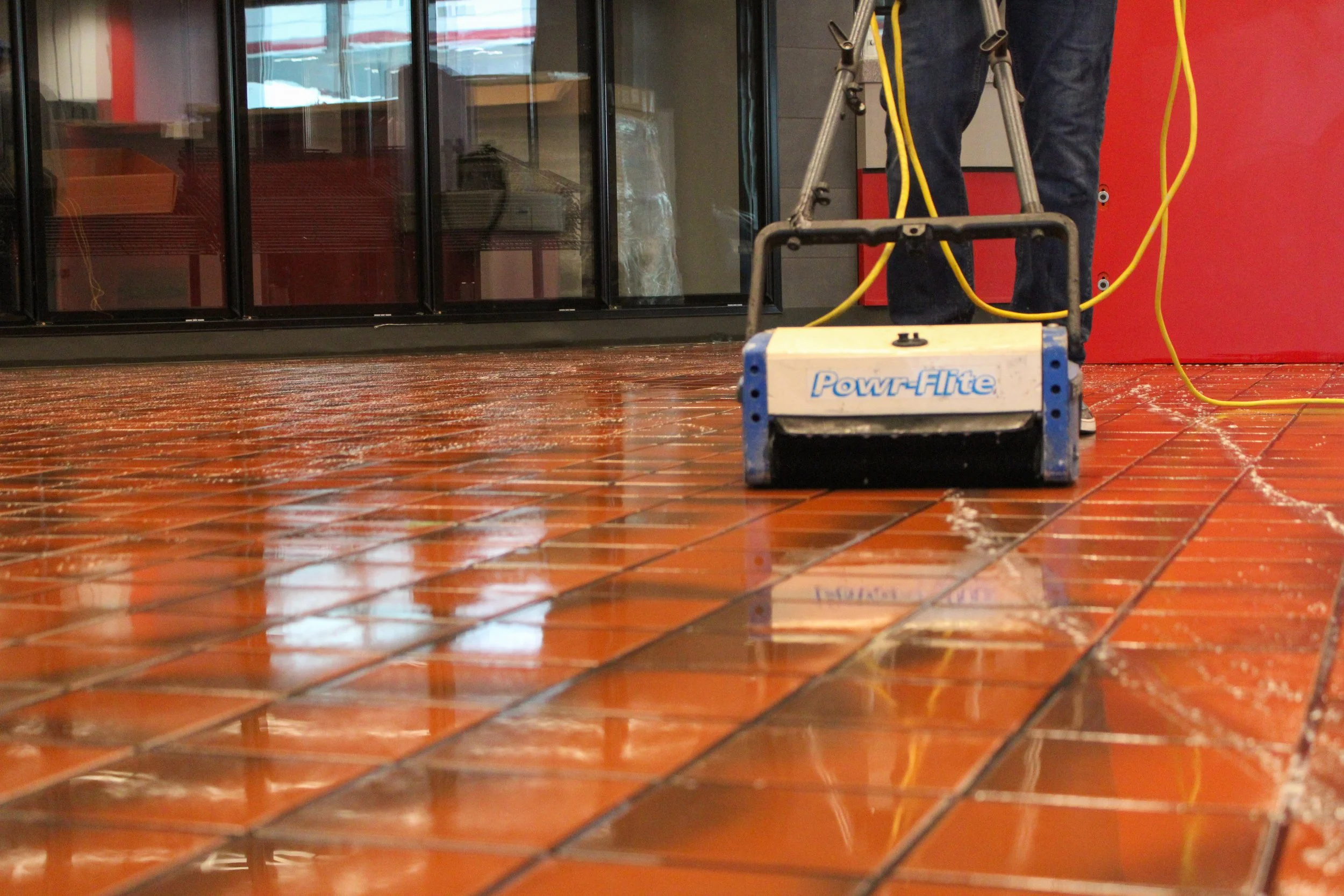A person in jeans standing on a polished tile floor using a Powr-Flite floor buffer with yellow cords passing through the stander's metal frame, next to a glass display case and a red wall.
