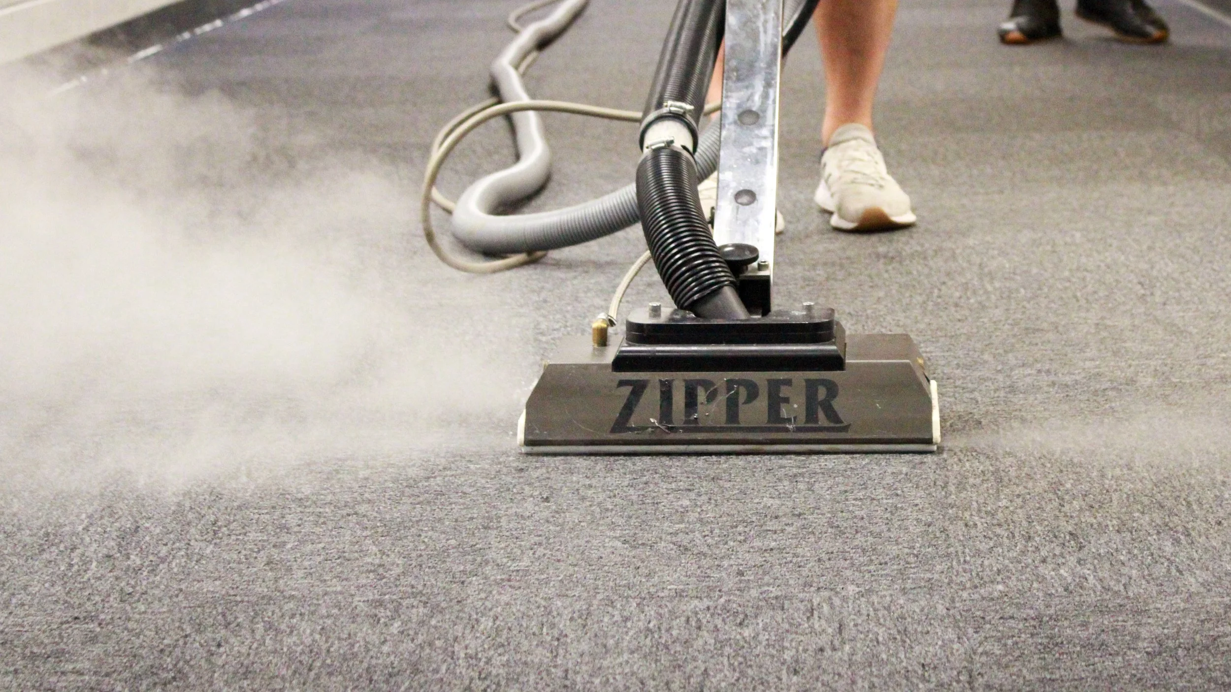 An industrial floor cleaning machine with the brand name 'ZIPPER' on the front, vacuuming a gray carpeted floor in a public space, with people walking in the background.