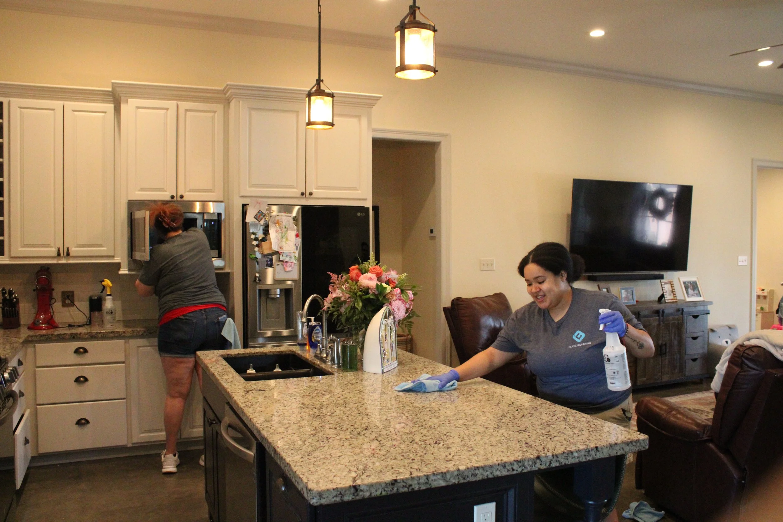 Inside a modern kitchen, two women are engaged in cleaning tasks. One woman with curly hair tied back, wearing a gray t-shirt and shorts, is reaching into an oven or microwave. The other woman, with dark hair pulled back, dressed in a blue-gray shirt, is smiling and spraying a cleaning solution on a counter, wearing gloves. The kitchen features white cabinets, a granite countertop, a bouquet of pink flowers on the counter, and a large flat-screen TV mounted on the wall in the background.