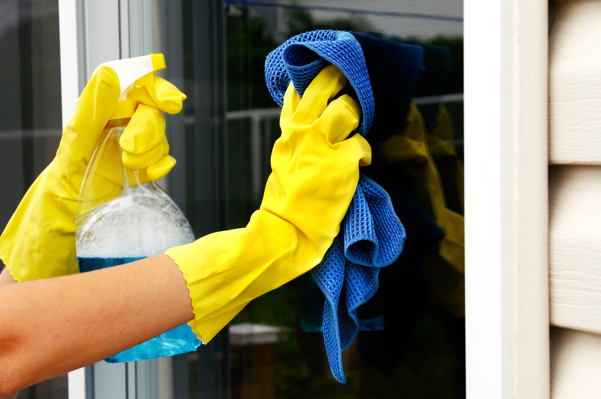 A person cleaning a glass window with a yellow spray bottle, wearing yellow rubber gloves and holding a blue cloth.
