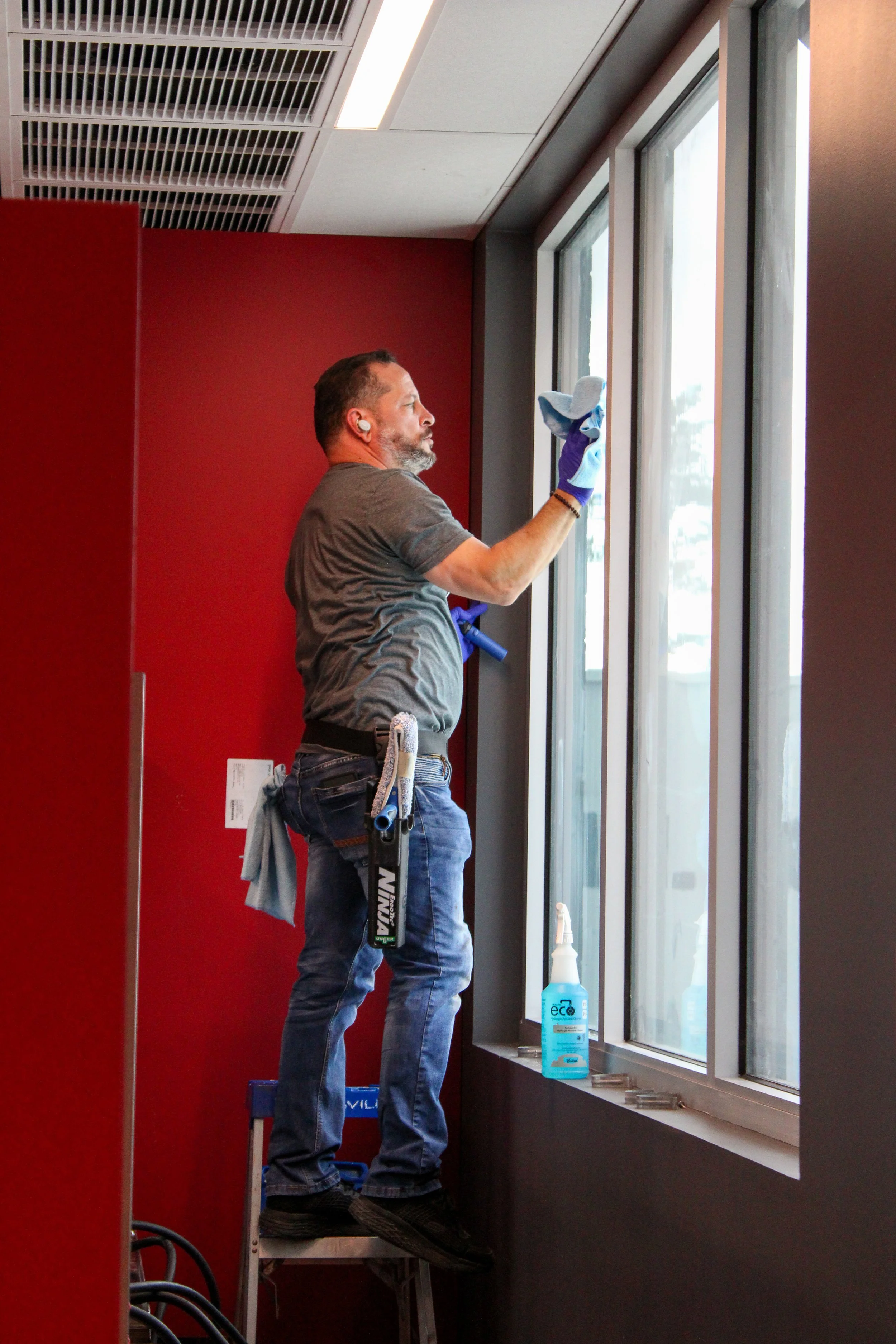 A man cleaning a window with a spray bottle and cloth inside a room with red and gray walls, and a ceiling with vents.