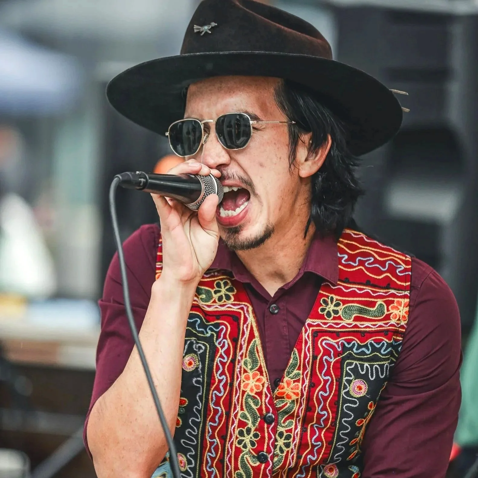 Singer in a wide-brimmed hat and embroidered shirt performing into a microphone at a Gio Chamba concert in Denver.