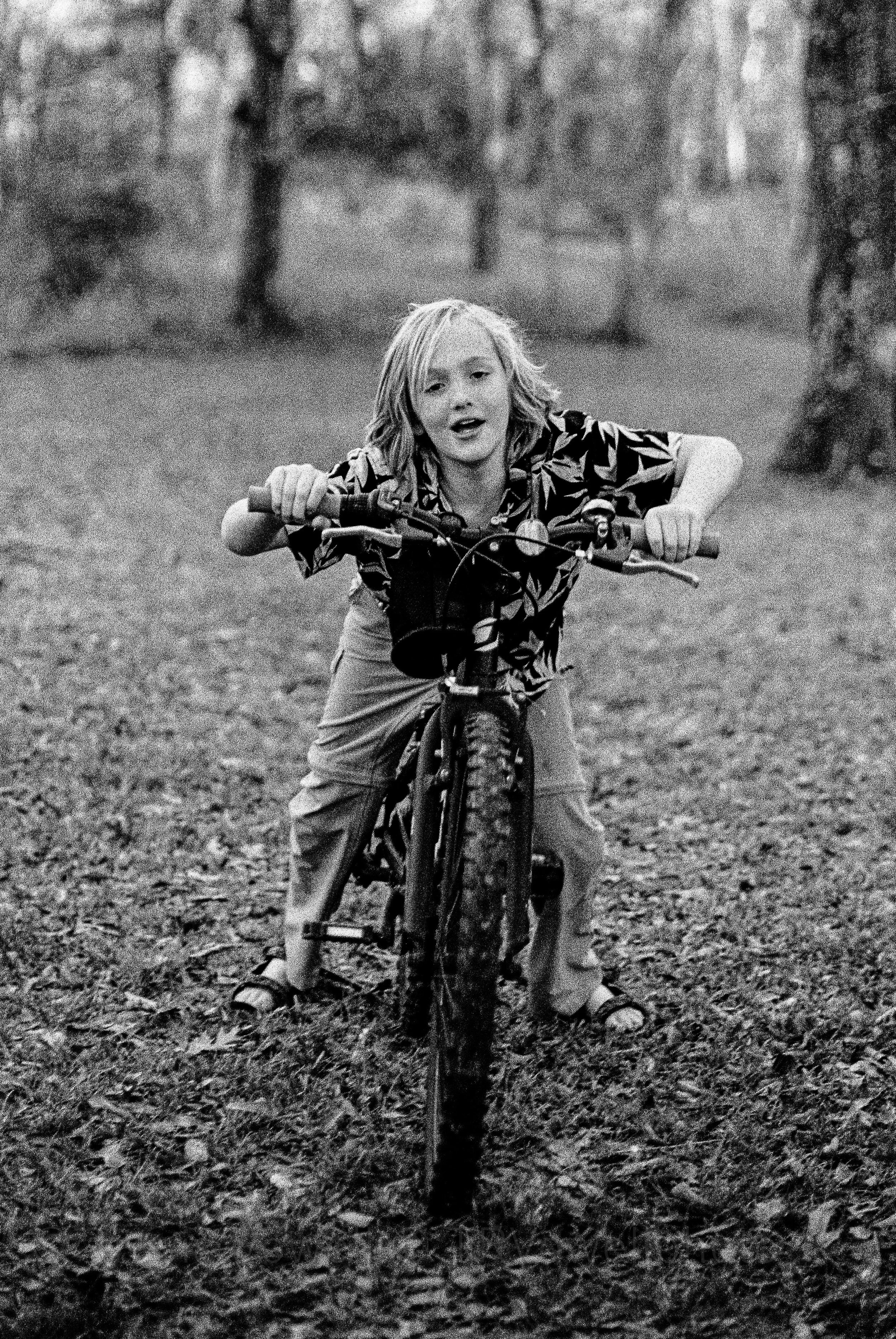 A young girl with shoulder-length hair riding a bicycle on a leaf-covered ground in a forested area.  Classic traditional old school retro Black and White B&W BW 35mm film photography shot on classic cameras real photos for history and keepsake docum