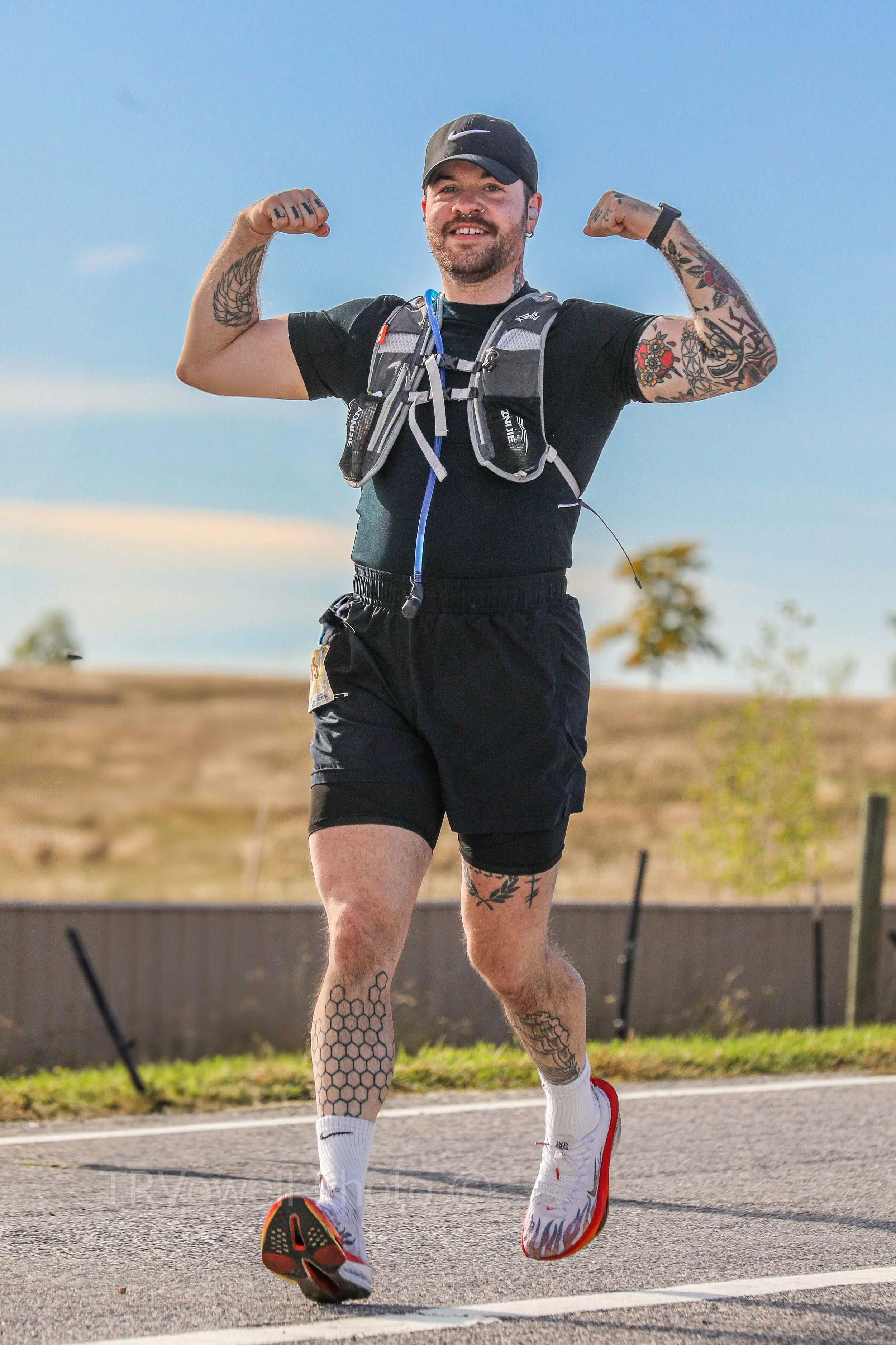 A man with tattoos on his arms and legs, wearing a black t-shirt, shorts, a black cap, and white running shoes, is seen running outdoors during daytime with a backpack.