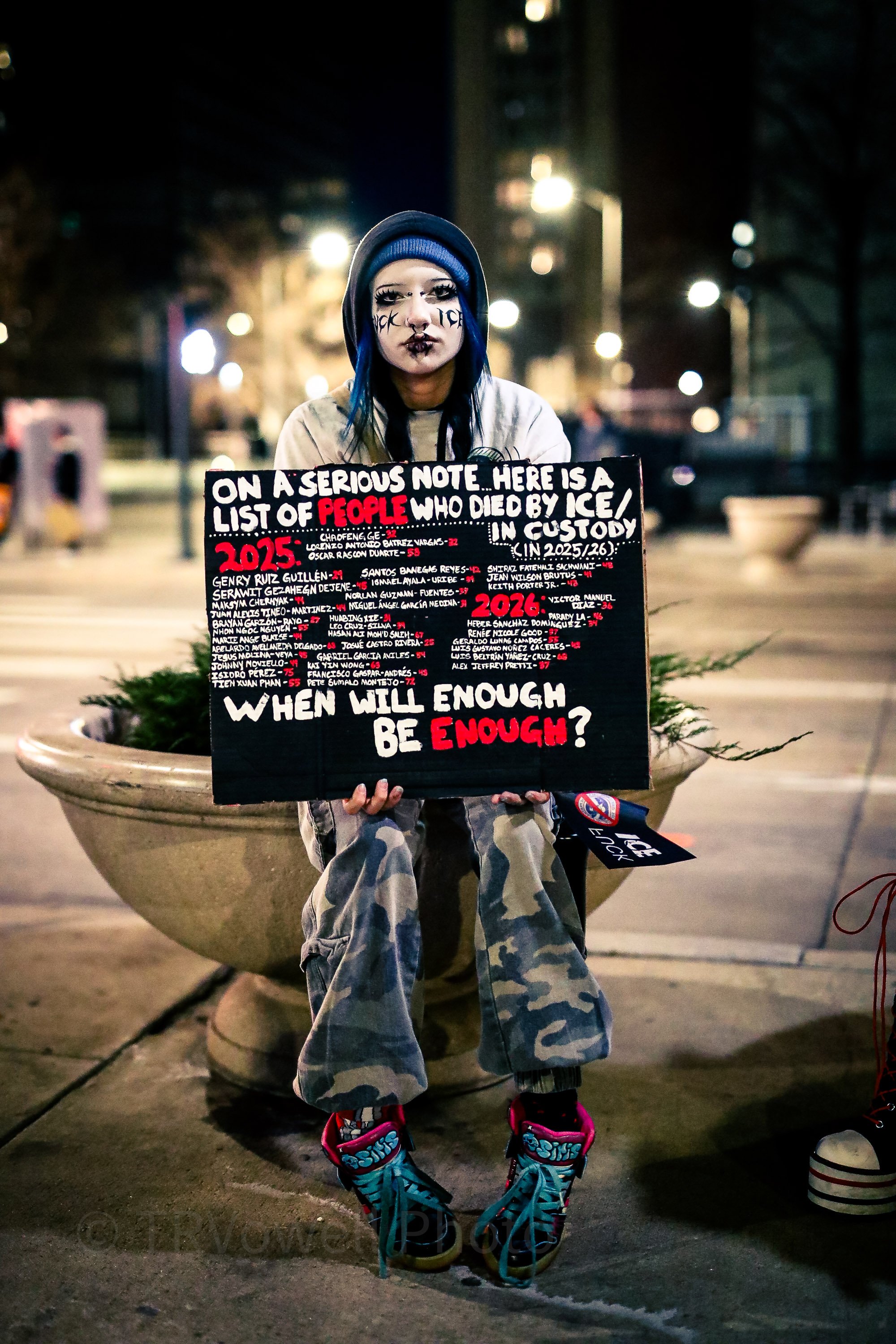 Participant at a nighttime ICE Out protest in Denver holds a sign listing deaths in ICE custody and asking, "When will enough be enough?"