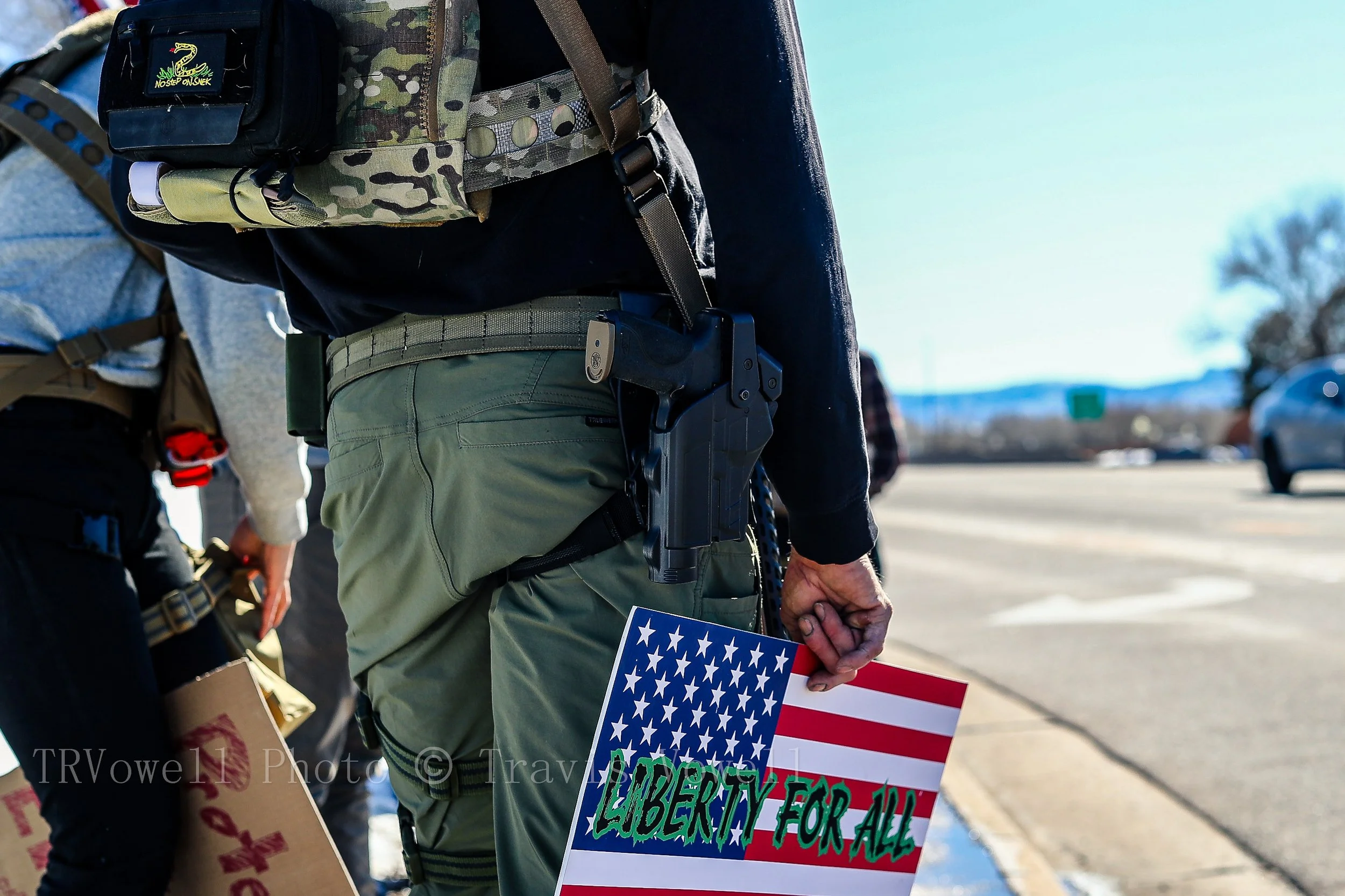 Close-up of tactical gear, a slung rifle, and a protest sign in Littleton, Colorado.