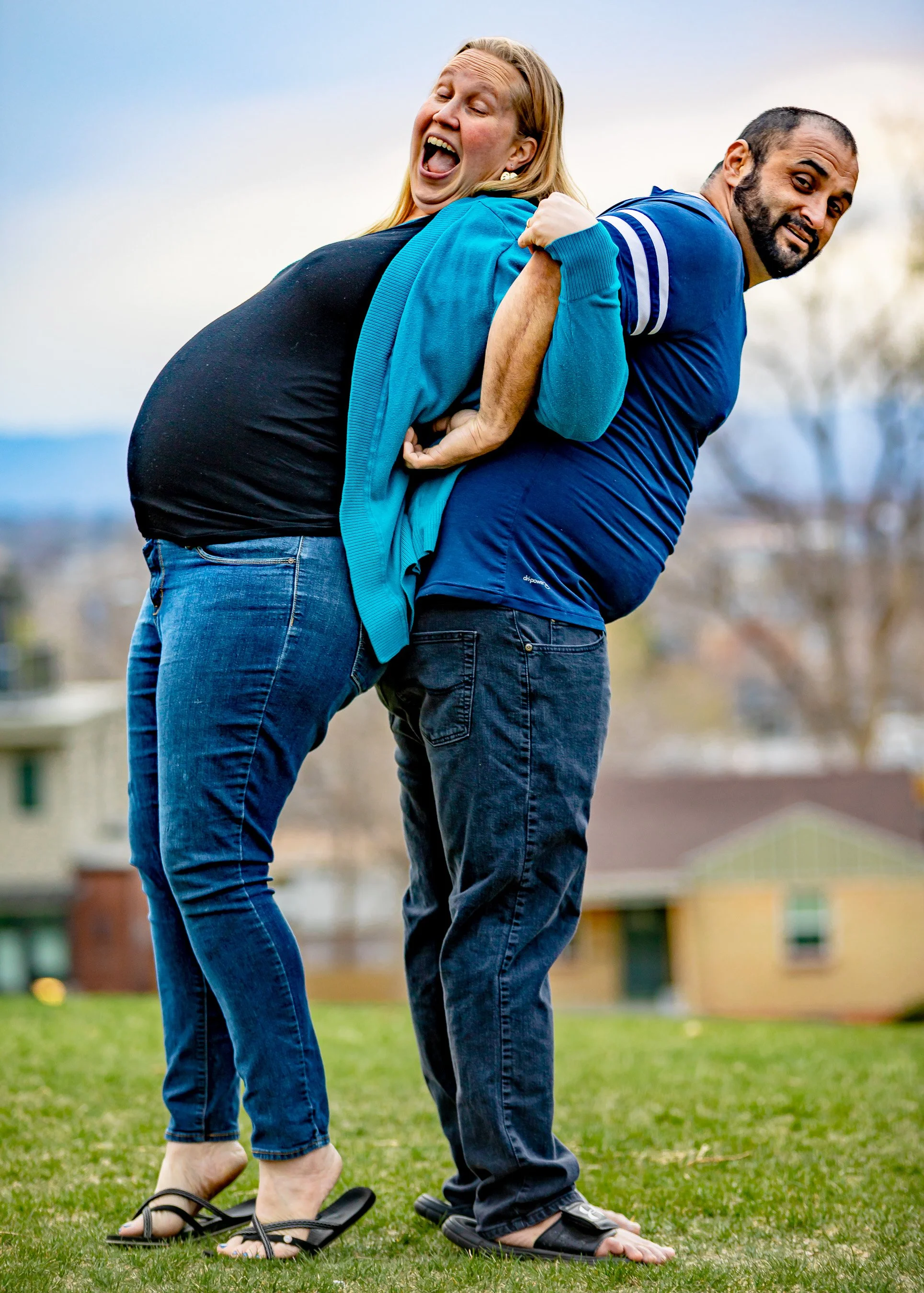 Two people, a woman and a man, are playfully pretending to be on a tandem bicycle with their legs and feet touching. They are standing on a grassy area outdoors with buildings and trees in the background, both wearing casual clothing and flip-flops. 