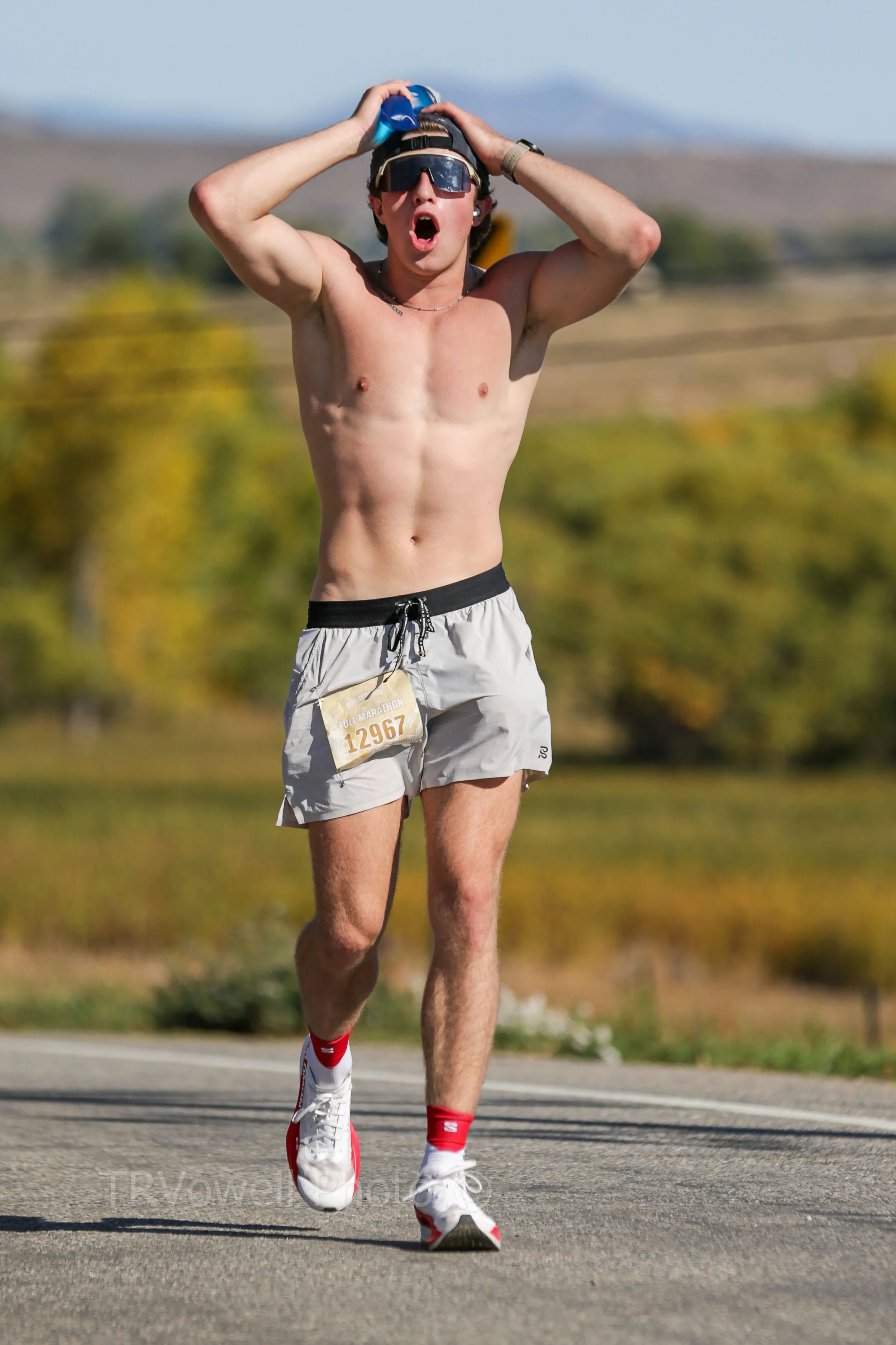 A shirtless male runner wearing sunglasses and a cap, holding his head with both hands, while running on a paved road during a marathon event with a bib number 12967.