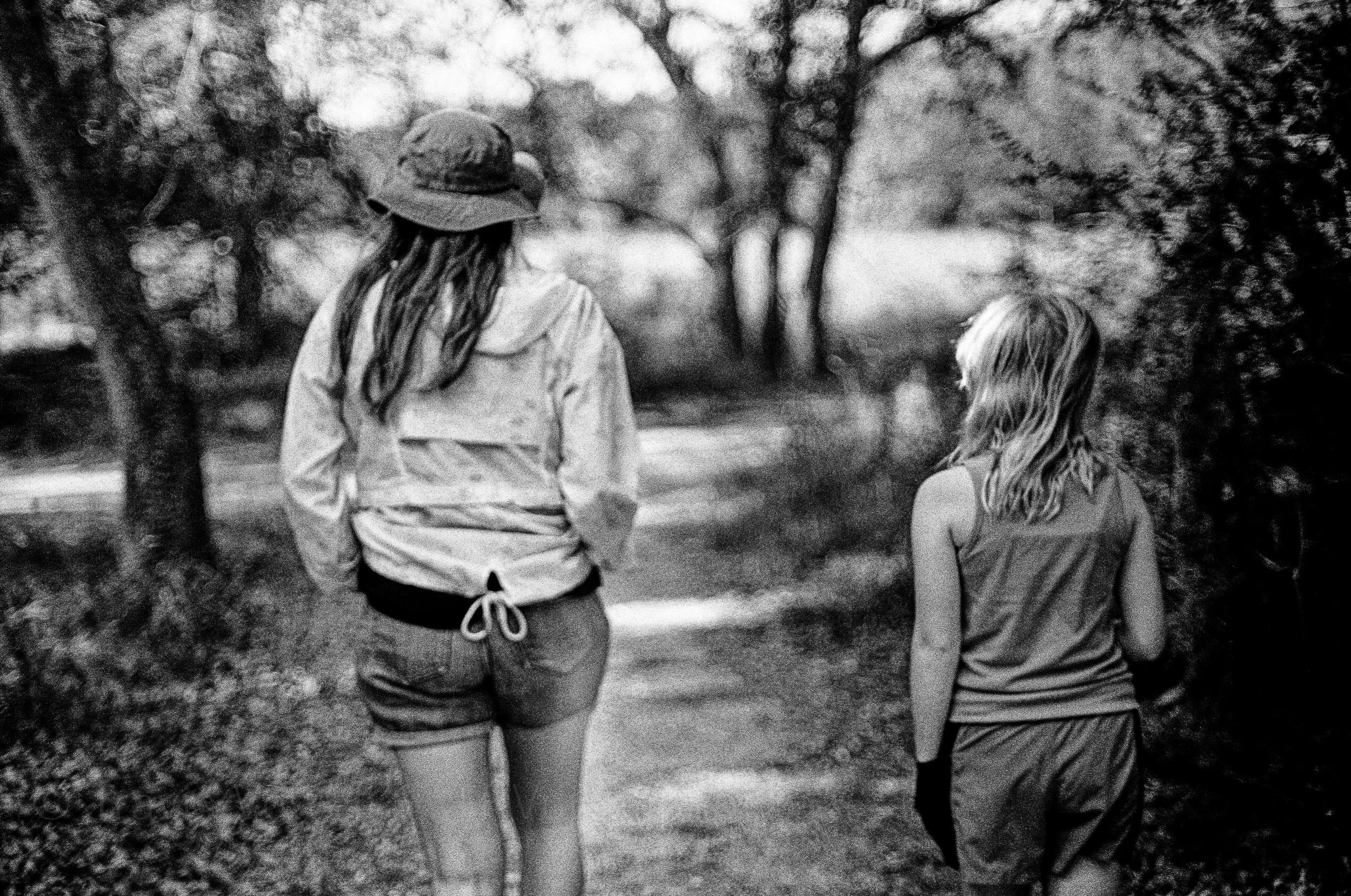 A black and white photo of two women walking on a path in a park, surrounded by trees and foliage, seen from behind.  Classic traditional old school retro Black and White B&W BW 35mm film photography shot on classic cameras real photos for history an