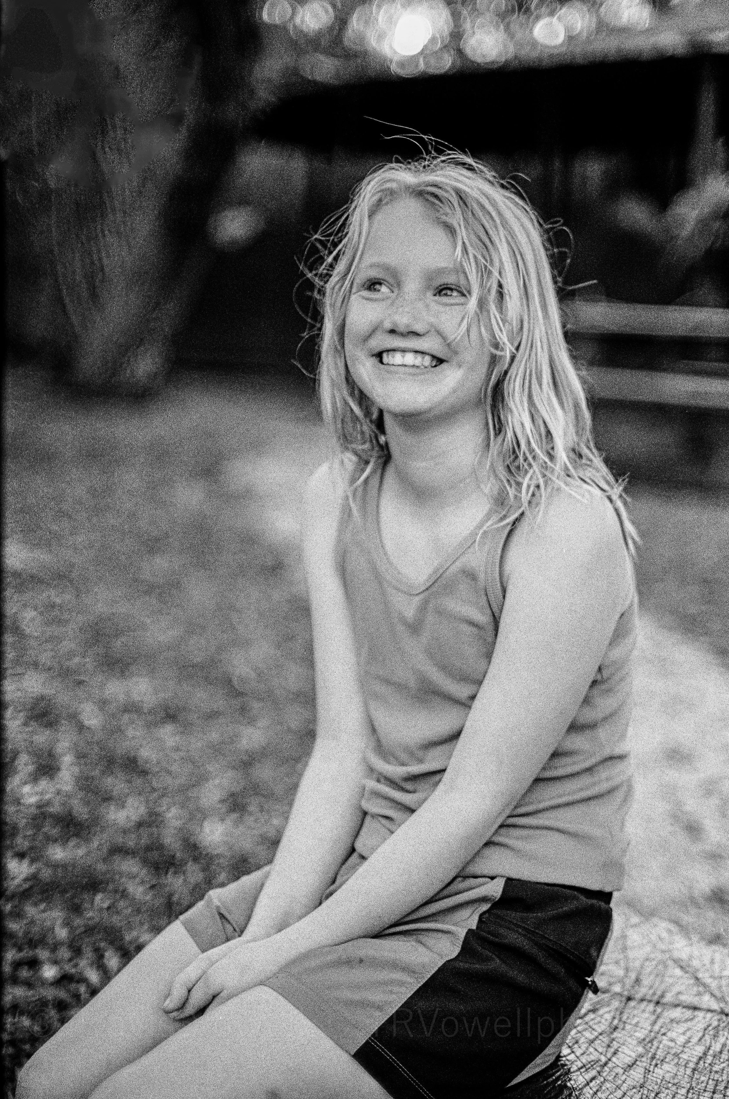 Portrait photo A young woman with curly blonde hair smiling and sitting outdoors near water, wearing a sleeveless top and shorts.  Classic traditional old school retro Black and White B&W BW 35mm film photography shot on classic cameras real photos f
