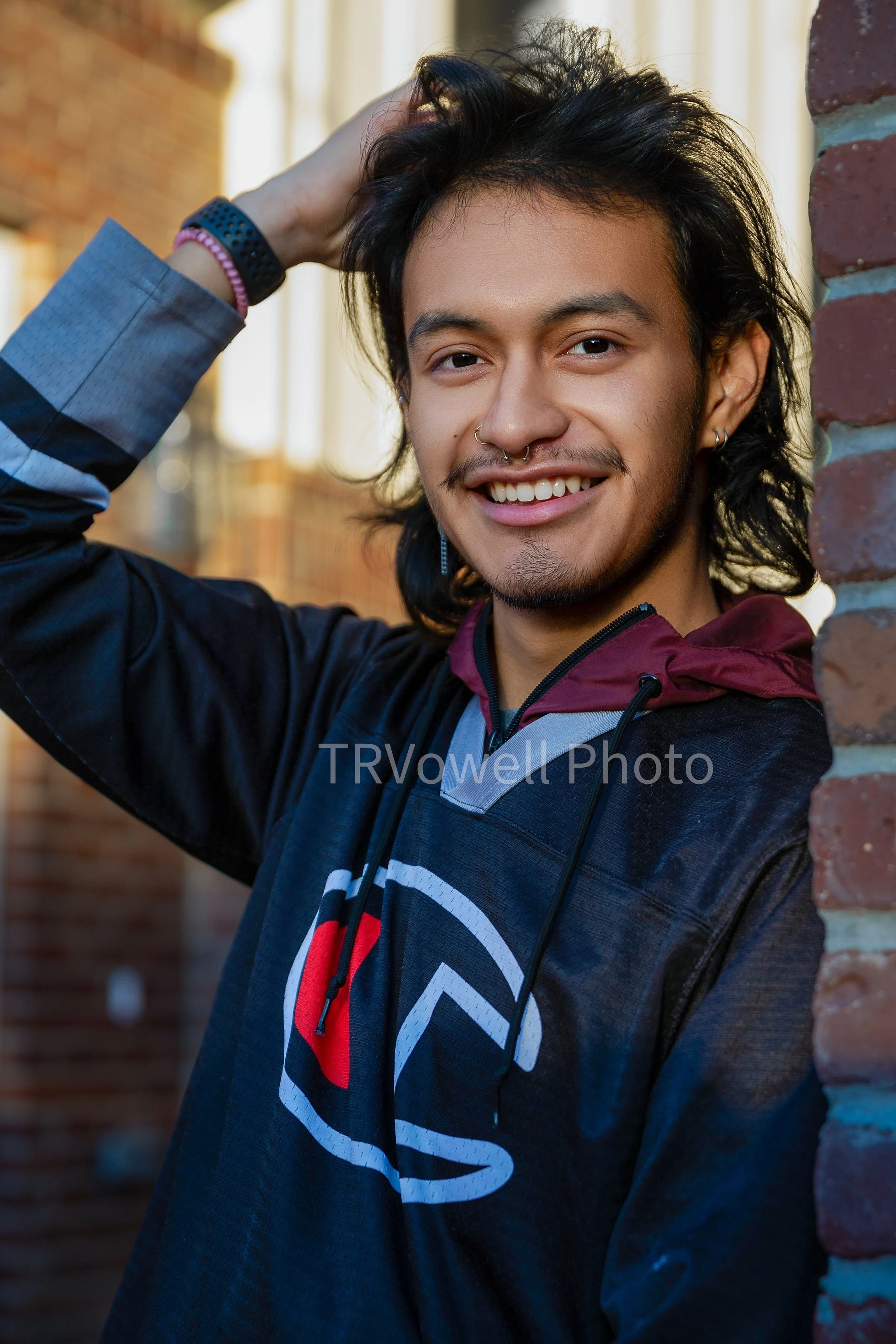 A young man with long, dark hair, wearing a black and red jacket, smiling and touching his hair, standing outside near a brick wall.