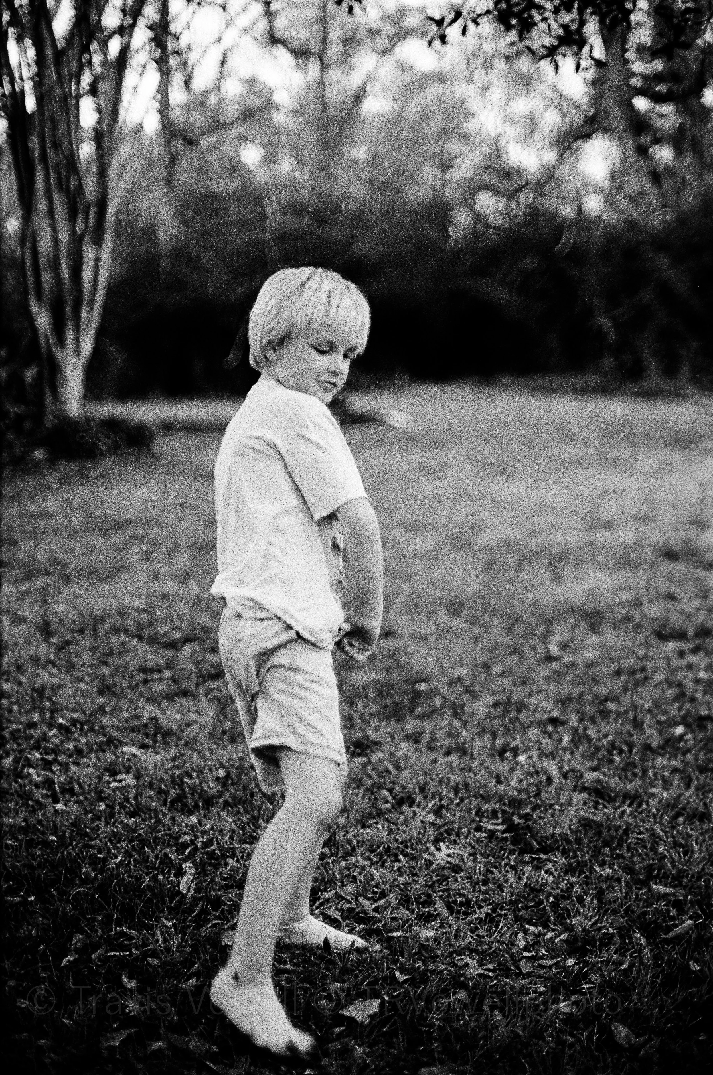 Classic portrait Black and White photo of A young boy with blonde hair wearing a t-shirt and shorts standing outdoors in a grassy area surrounded by trees, captured in black and white.  Classic traditional old school retro Black and White B&W BW 35mm