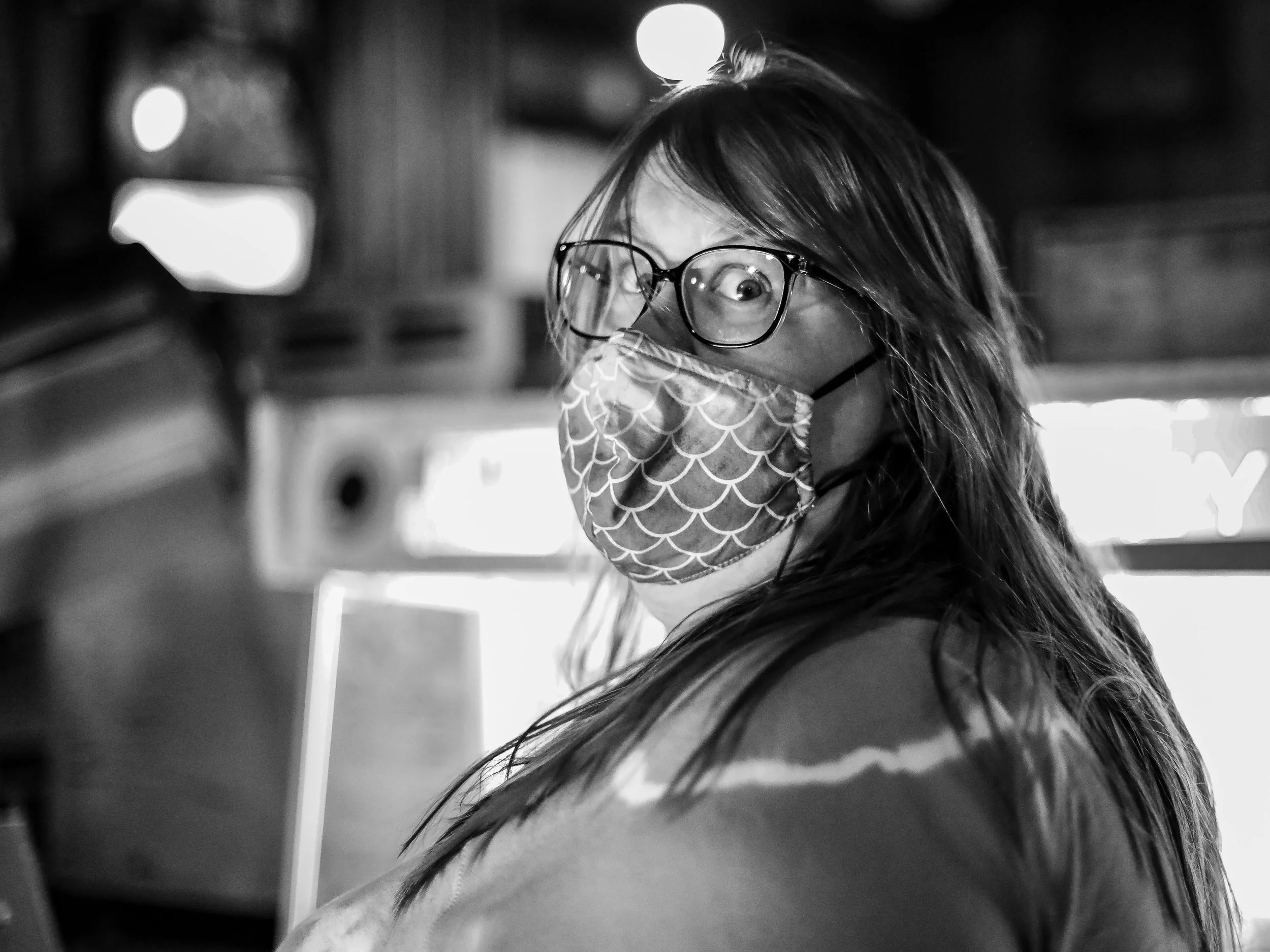 Black and white photo of a woman with long hair wearing glasses and a patterned face mask, looking over her shoulder with her eyes wide open in a surprised or curious expression.  Real memories and real emotions documentary photography style portrait
