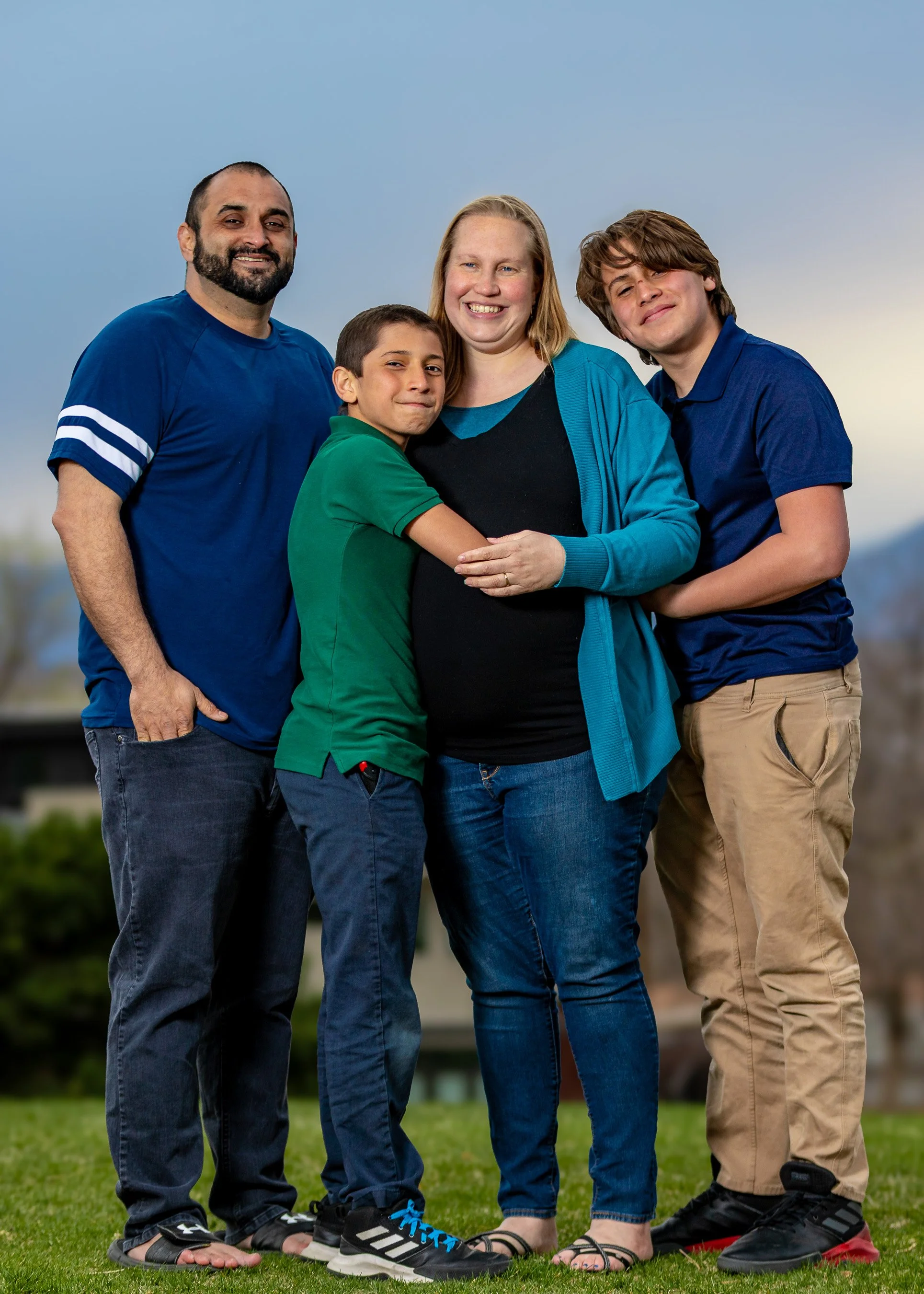 A diverse family of four standing outdoors on grass, smiling, with two adults and two children, dressed casually and standing close together.  Golden hour sunset portraits editorial style pics