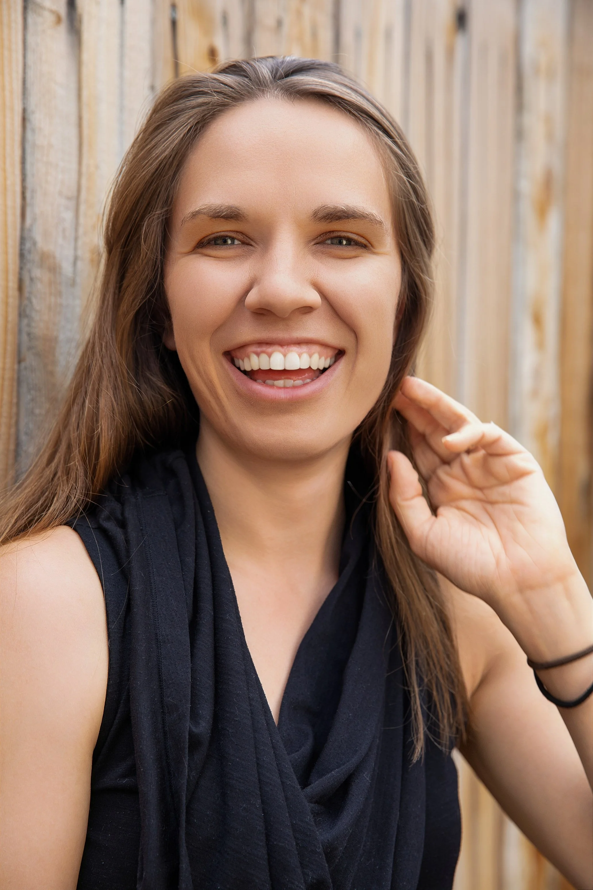 Portrait of A smiling young woman with long brown hair, wearing a black sleeveless top, standing in front of a wooden fence.  Dating profile email social marketing influencer outdoor