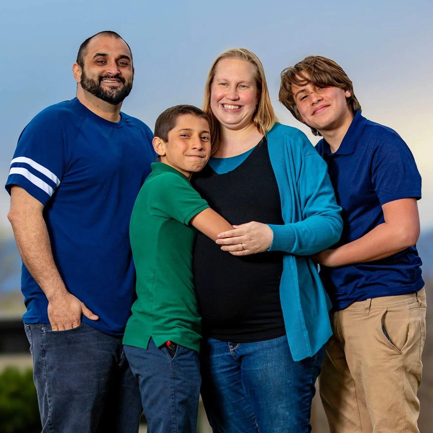 Outdoor family portrait of two adults and two children standing close together with their arms around one another.