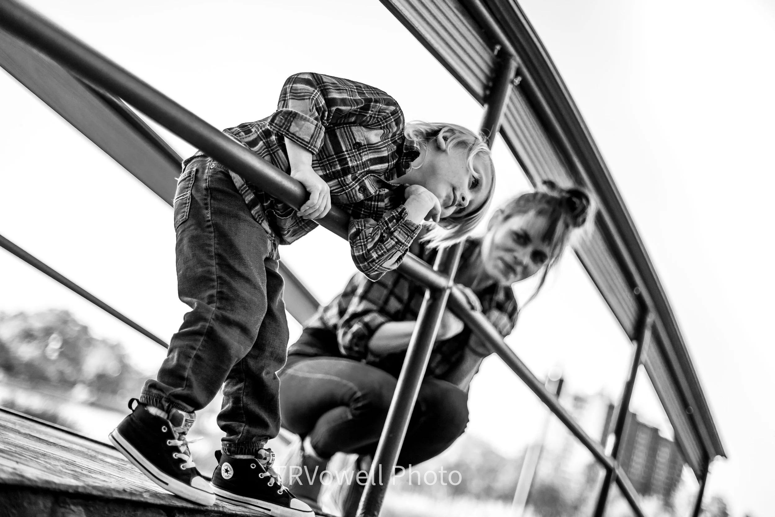 A young boy leaning over a railing at a playground, next to a woman sitting under the same structure, both looking downward. The photo is in black and white.