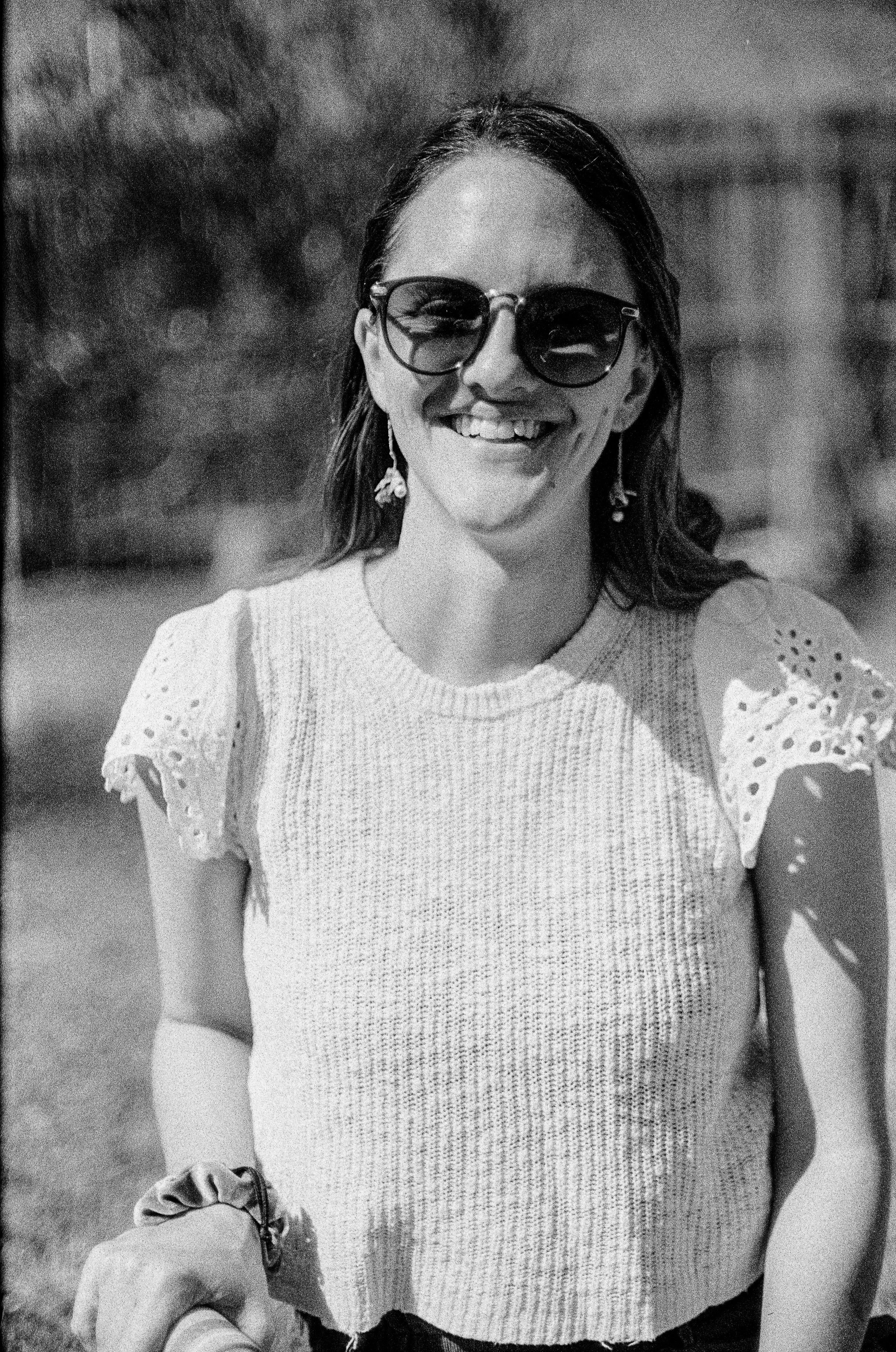 Black and white photo portrait of a woman wearing sunglasses and a patterned blouse, smiling outdoors.  Classic traditional old school retro Black and White B&W BW 35mm film photography shot on classic cameras real photos for history and keepsake doc