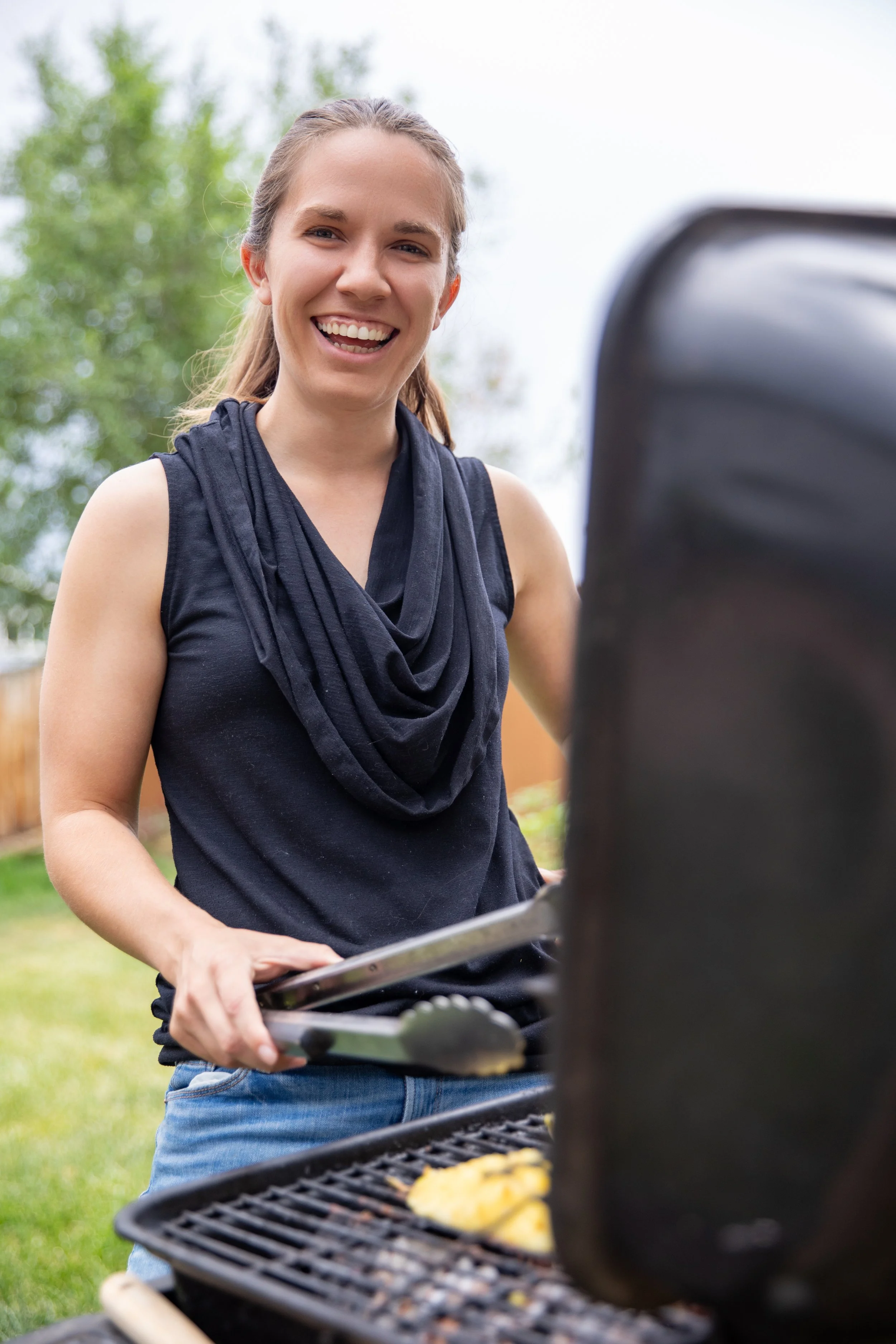 A woman outdoors grilling healthy food on a barbecue, smiling at the camera.  Branding social media email photos